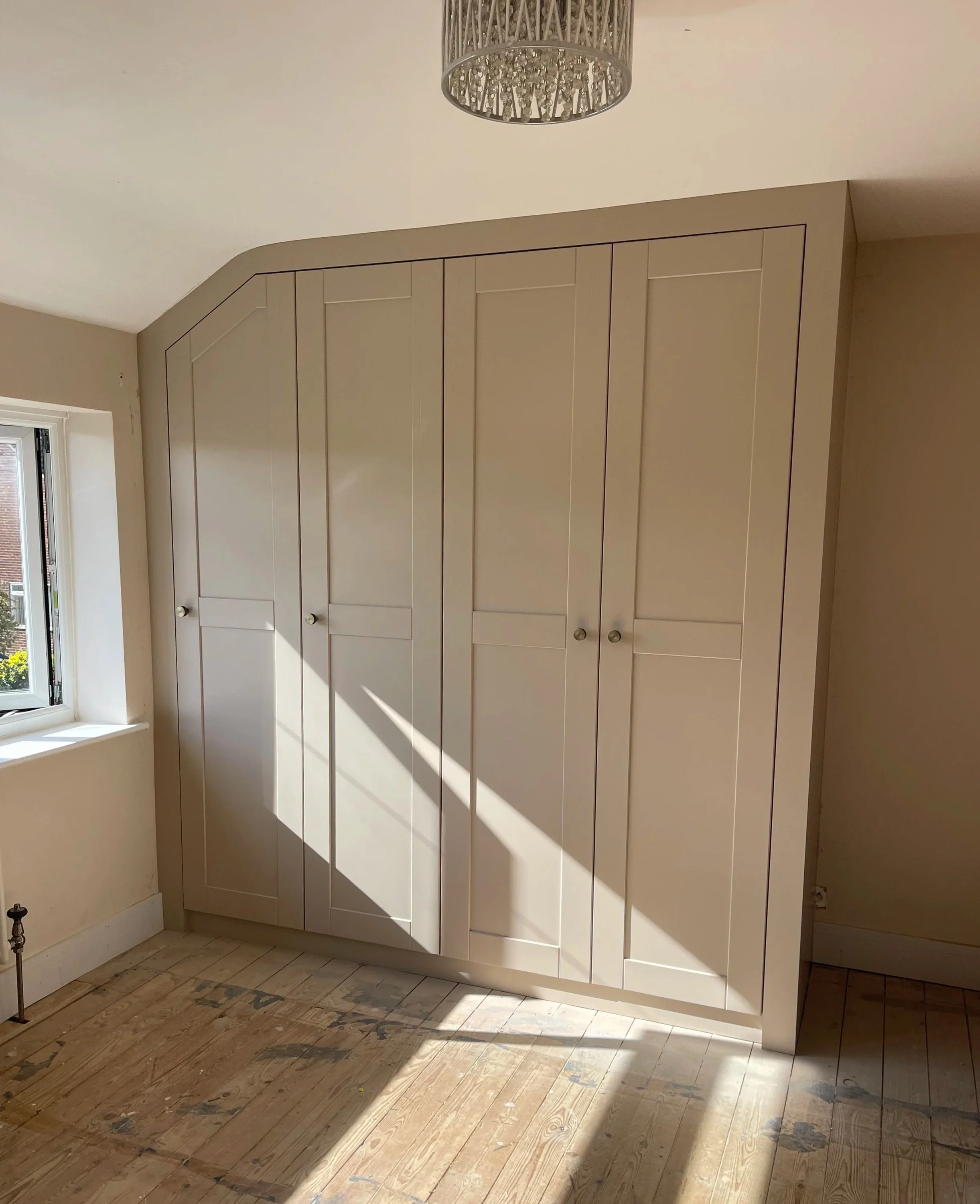 A white wardrobe with paneled doors in a room with a wooden floor and a window on the left, sunlight casting shadows on the wardrobe.