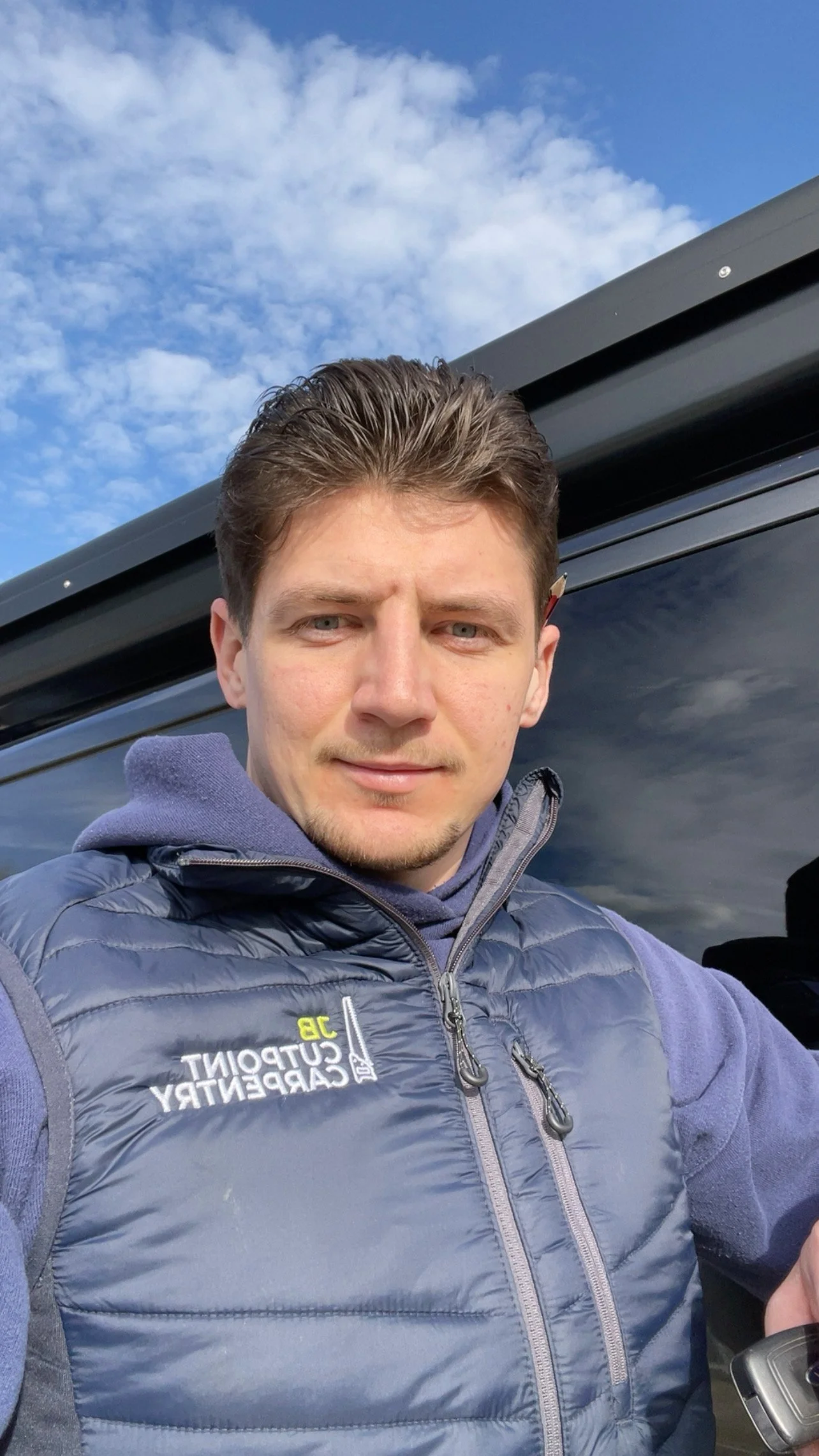 A young man with short brown hair wearing a blue jacket and purple hoodie, taking a selfie outdoors with a blue sky and some clouds and a black vehicle in the background.