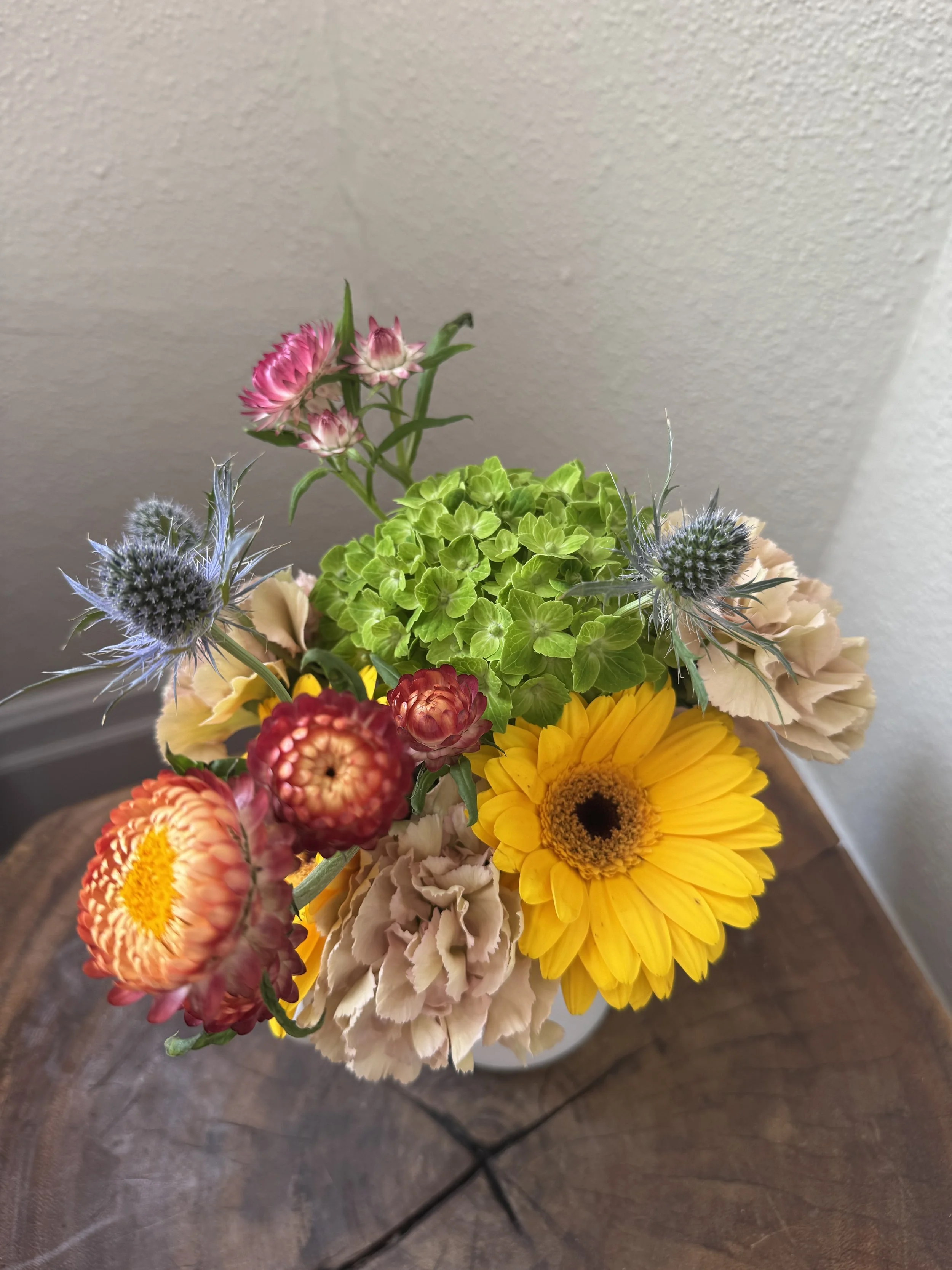 Colorful bouquet of various flowers including yellow gerbera daisy, pink and red small flowers, green hydrangea, and blue thistle in a white vase on a wooden table.
