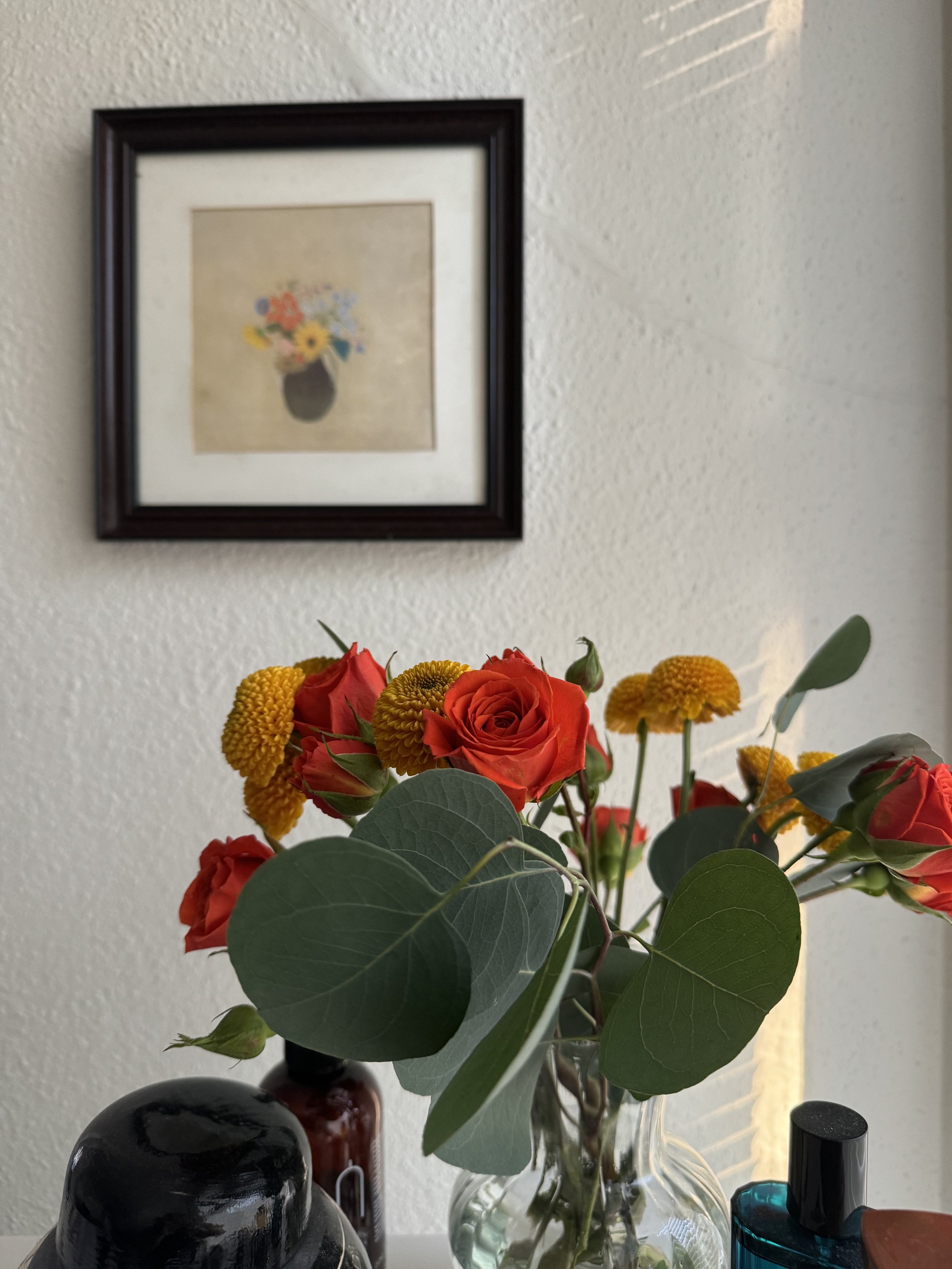 A clear glass vase with a bouquet of red roses, small yellow flowers, and eucalyptus leaves placed on a white surface. Behind the vase, there are several bottles, including a black bottle and an amber bottle. A framed picture of a flower arrangement hangs on a white textured wall above the bouquet.