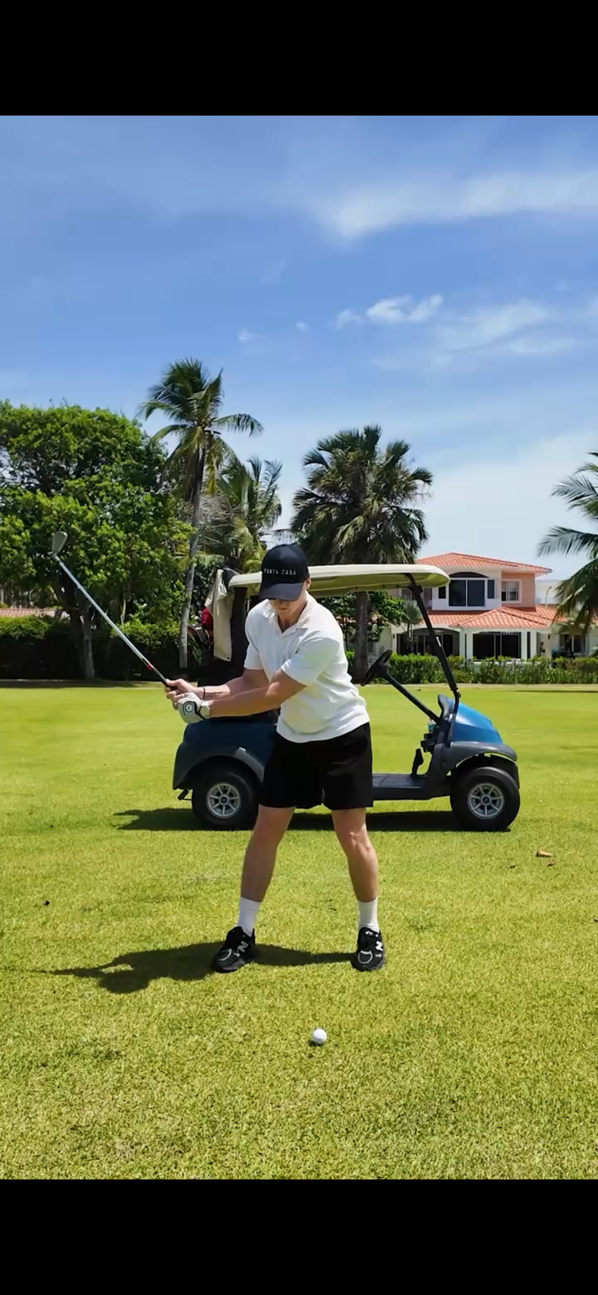 Person preparing to hit golf ball on lush green golf course with trees, a golf cart, and houses in the background under a blue sky with a few clouds.