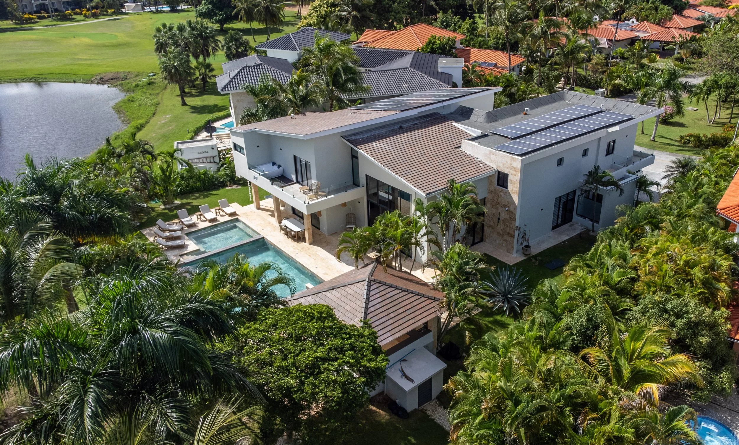 Aerial view of a modern house with solar panels on the roof, a swimming pool, and lush tropical landscaping including palm trees, neighboring houses, a pond, and a golf course in the background.