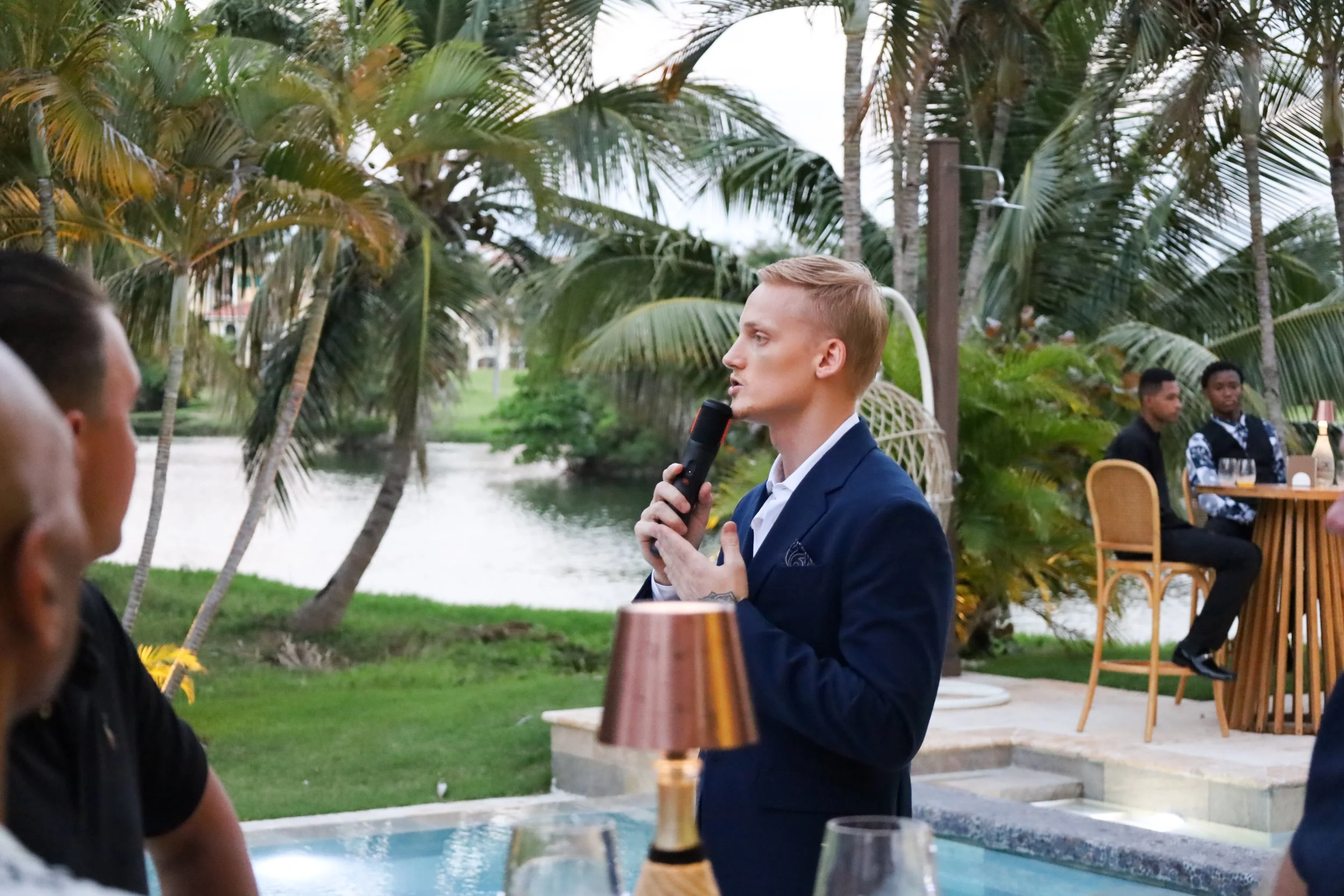A man in a blue suit holding a microphone, speaking outdoors near a swimming pool with tropical palm trees around, during a social gathering or event at sunset or early evening.