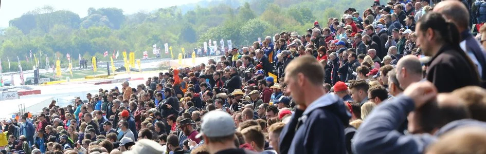 Crowd of people attending an outdoor event, seated on tiered bleachers, with a race track and flags in the background.