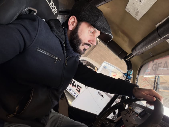 A man with a beard wearing a flat cap, black hoodie, and black jacket, sitting in the driver's seat of a vehicle, looking to the right, with a dashboard and interior parts visible.