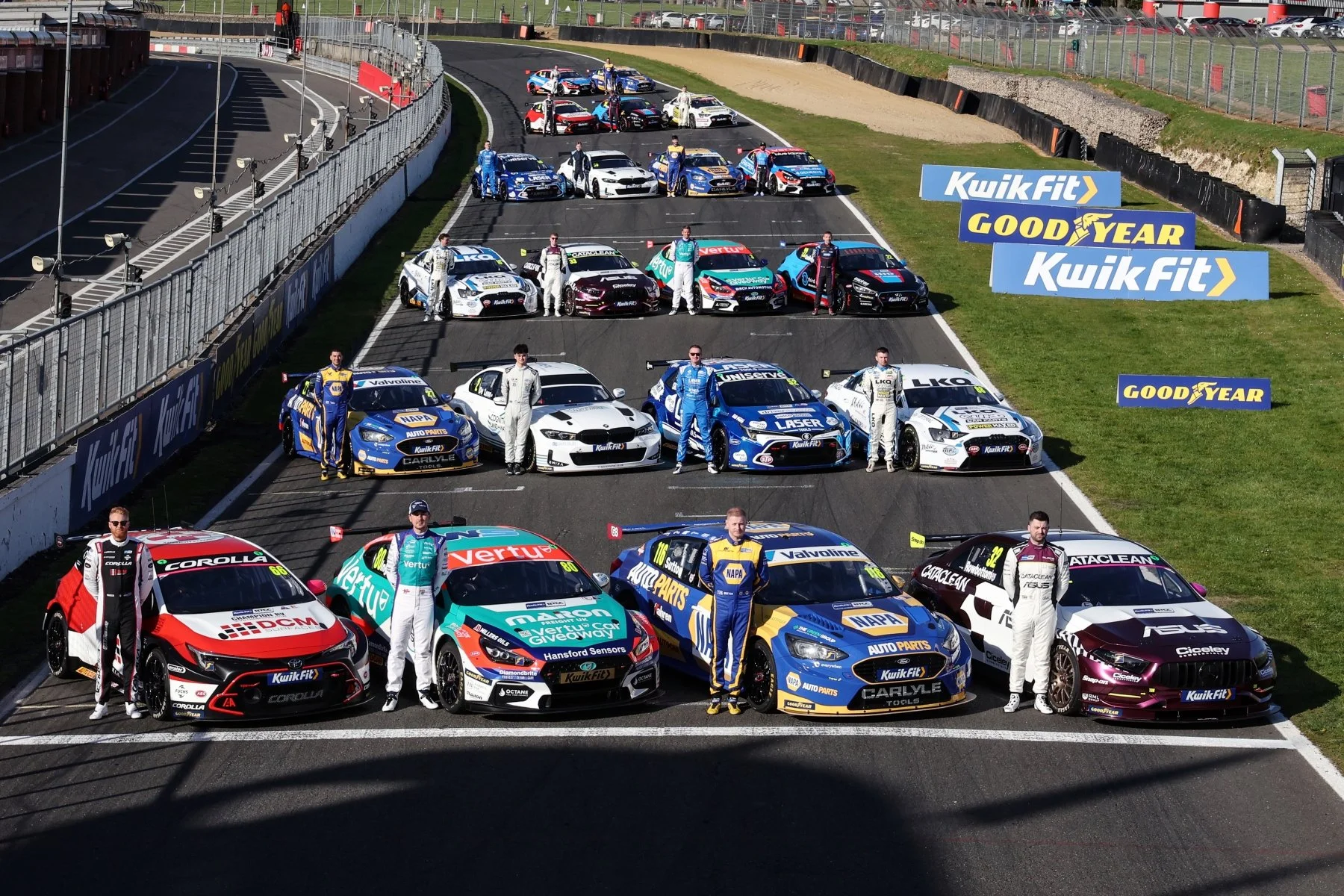 Race cars and drivers lined up on a racetrack ahead of a race, with sponsor banners along the side.