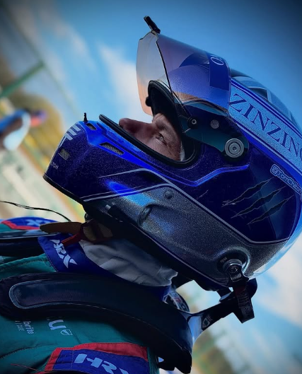 Person wearing a blue racing helmet and racing suit, looking to the right, with a cloudy sky in the background.