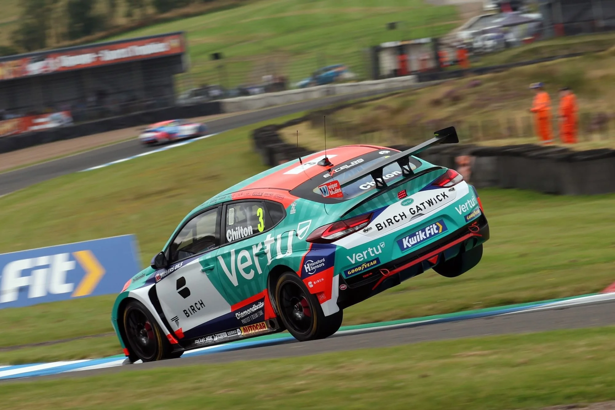 A race car on a track lifting off the ground, with a second car in the background, surrounded by grass, asphalt, and tire barriers.