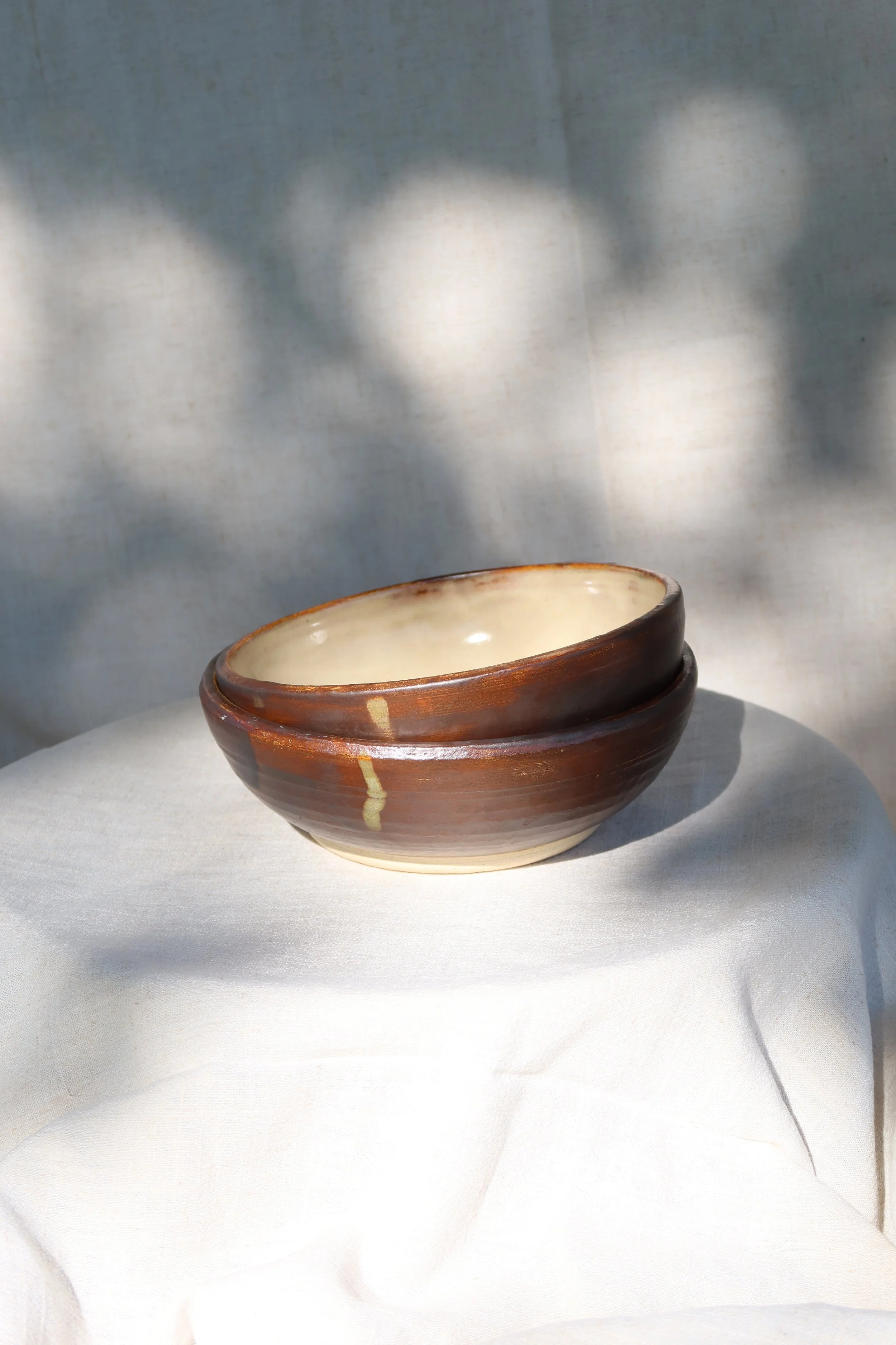 Two stacked ceramic bowls with a rustic brown and cream glaze, placed on a white cloth with soft natural light and shadows.