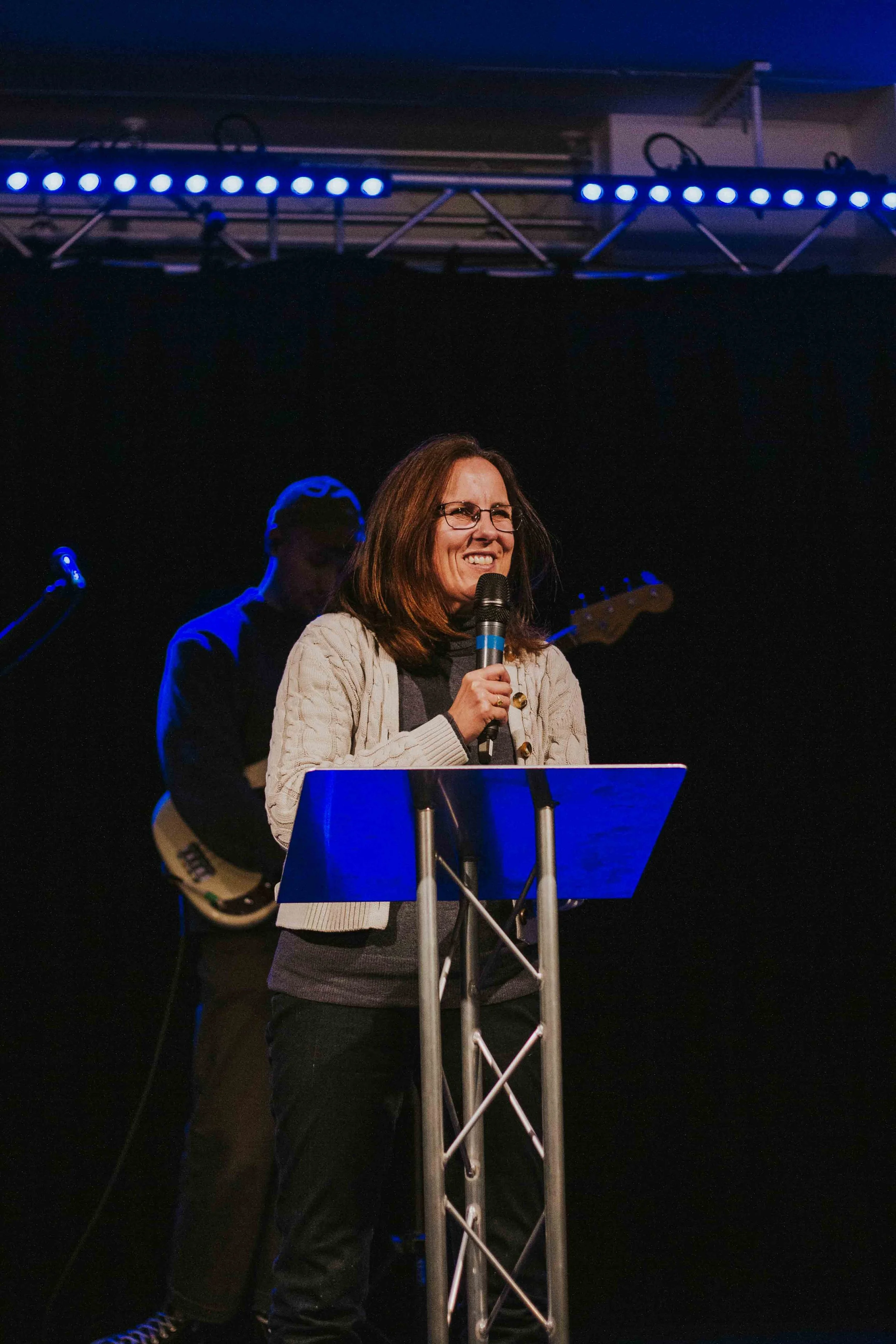 A woman with brown hair and glasses is smiling while speaking into a microphone at a podium on a stage. A man playing an electric guitar is in the background.