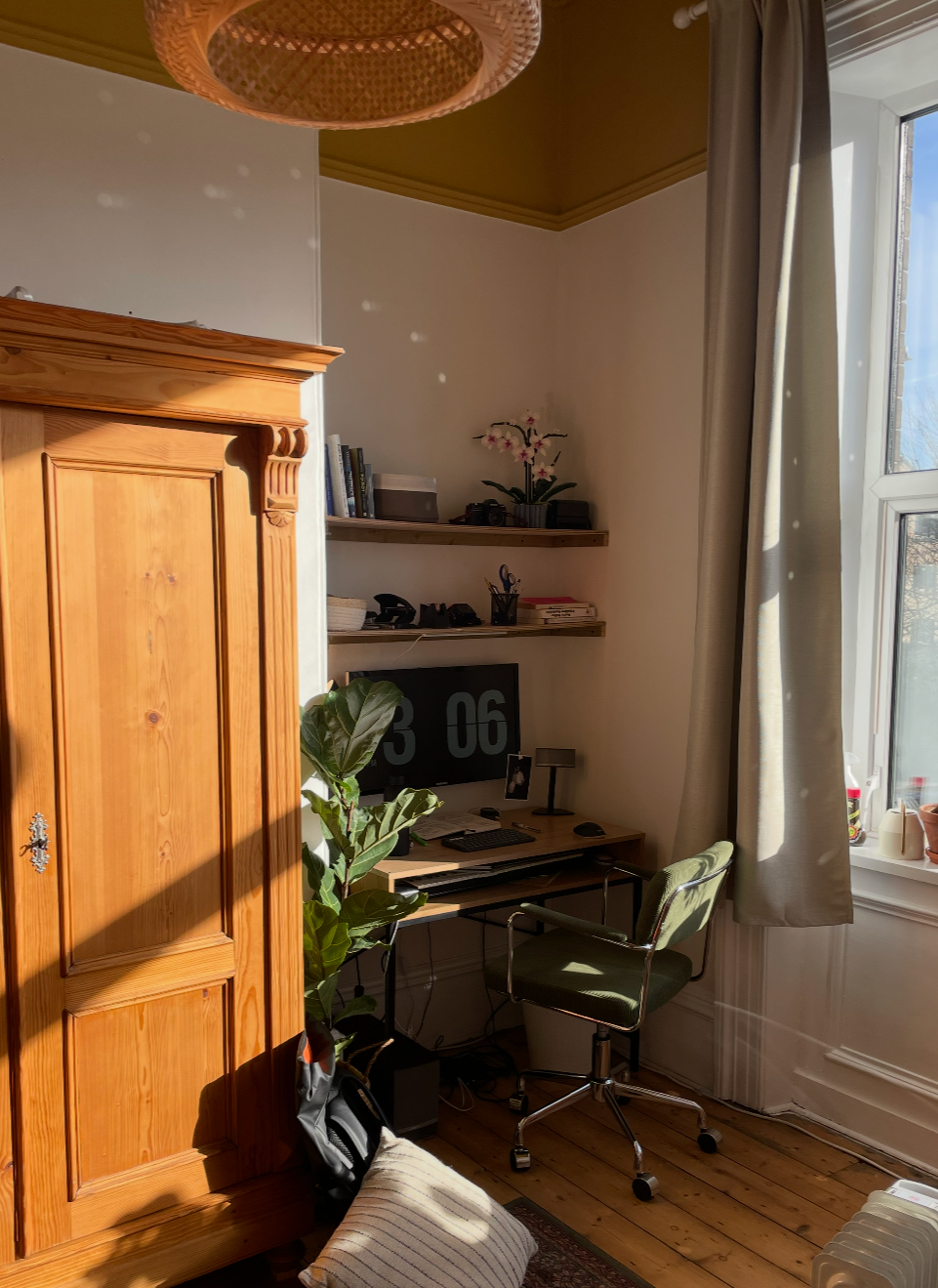 A corner of a room with a wooden wardrobe, a desk with a monitor, bookshelves, a green office chair, and a window with curtains letting in sunlight.