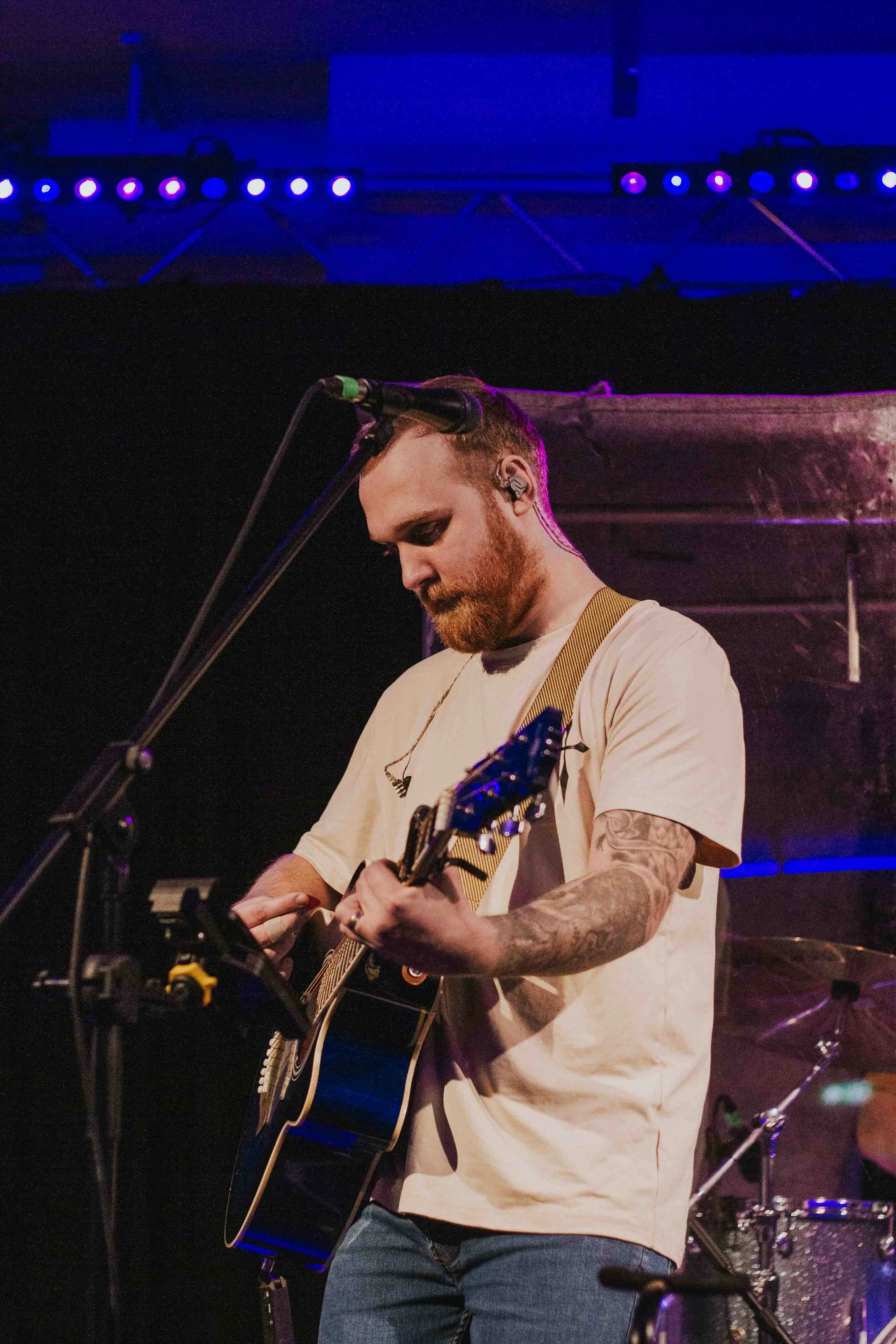 Male musician playing an acoustic guitar on stage with a microphone, stage lights, and drums in the background.