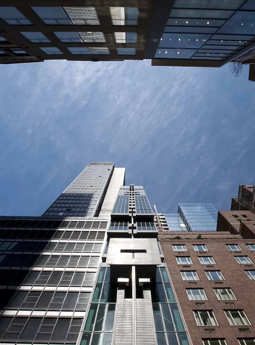 View of tall modern skyscrapers from ground level, looking up at a clear blue sky with wispy clouds.