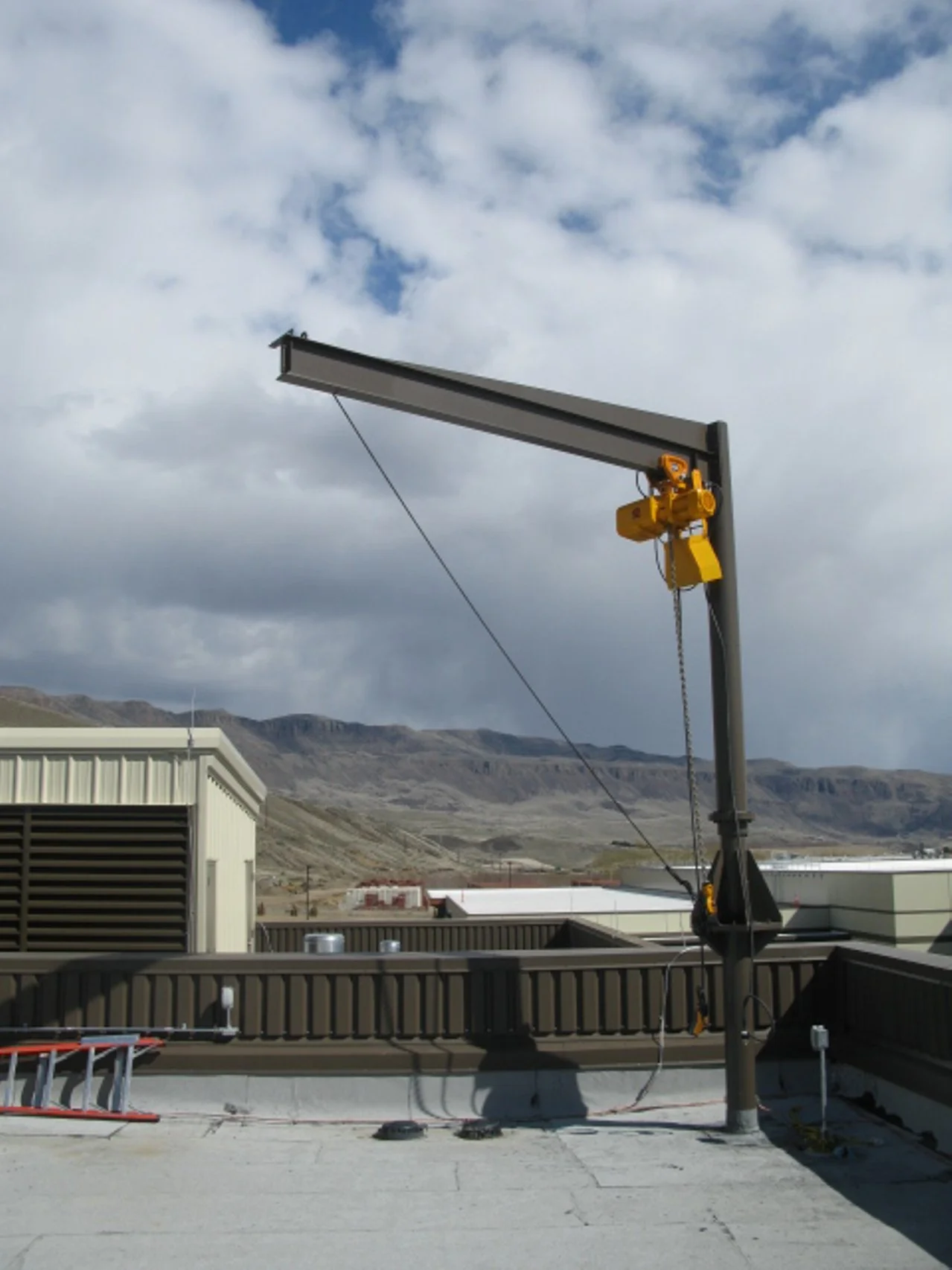 Industrial rooftop with a yellow crane, rooftop equipment, and a mountain landscape in the background under cloudy skies.
