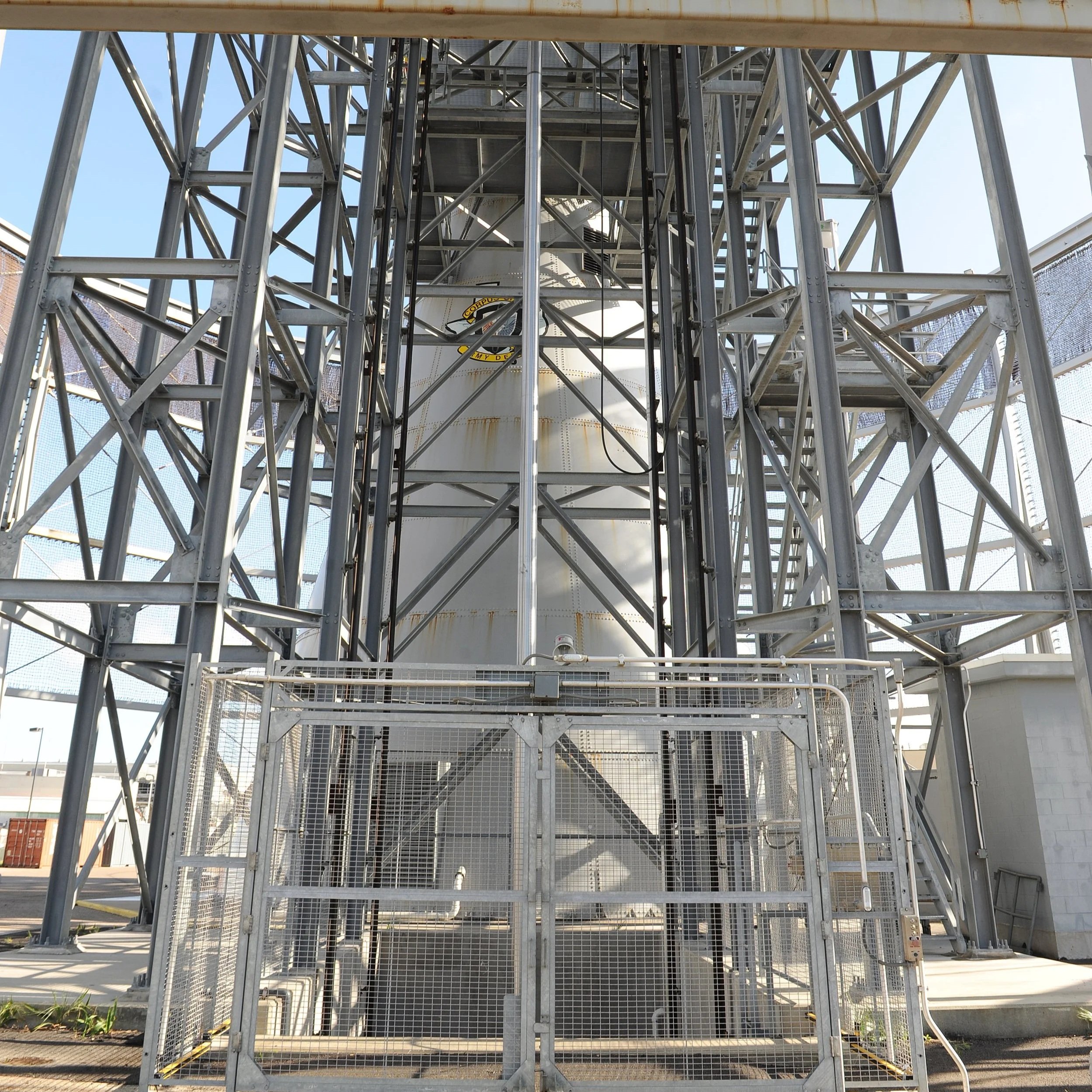 A metallic industrial structure with multiple levels, ladders, and railings, possibly part of a relay or communication station, set outdoors under a clear blue sky.