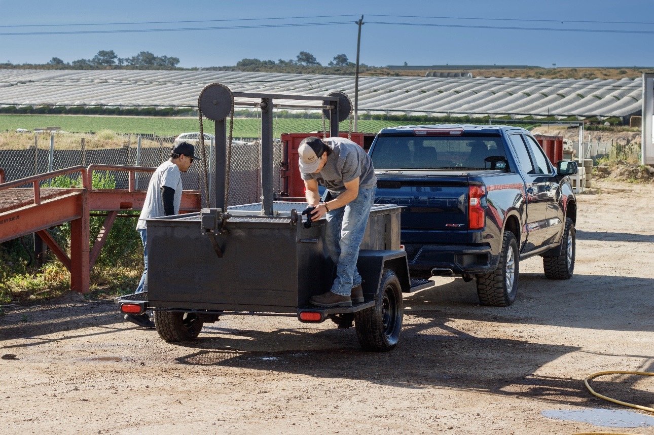 Two people working outdoors at a farmland or nursery, one on a trailer and the other nearby, with greenhouses in the background.