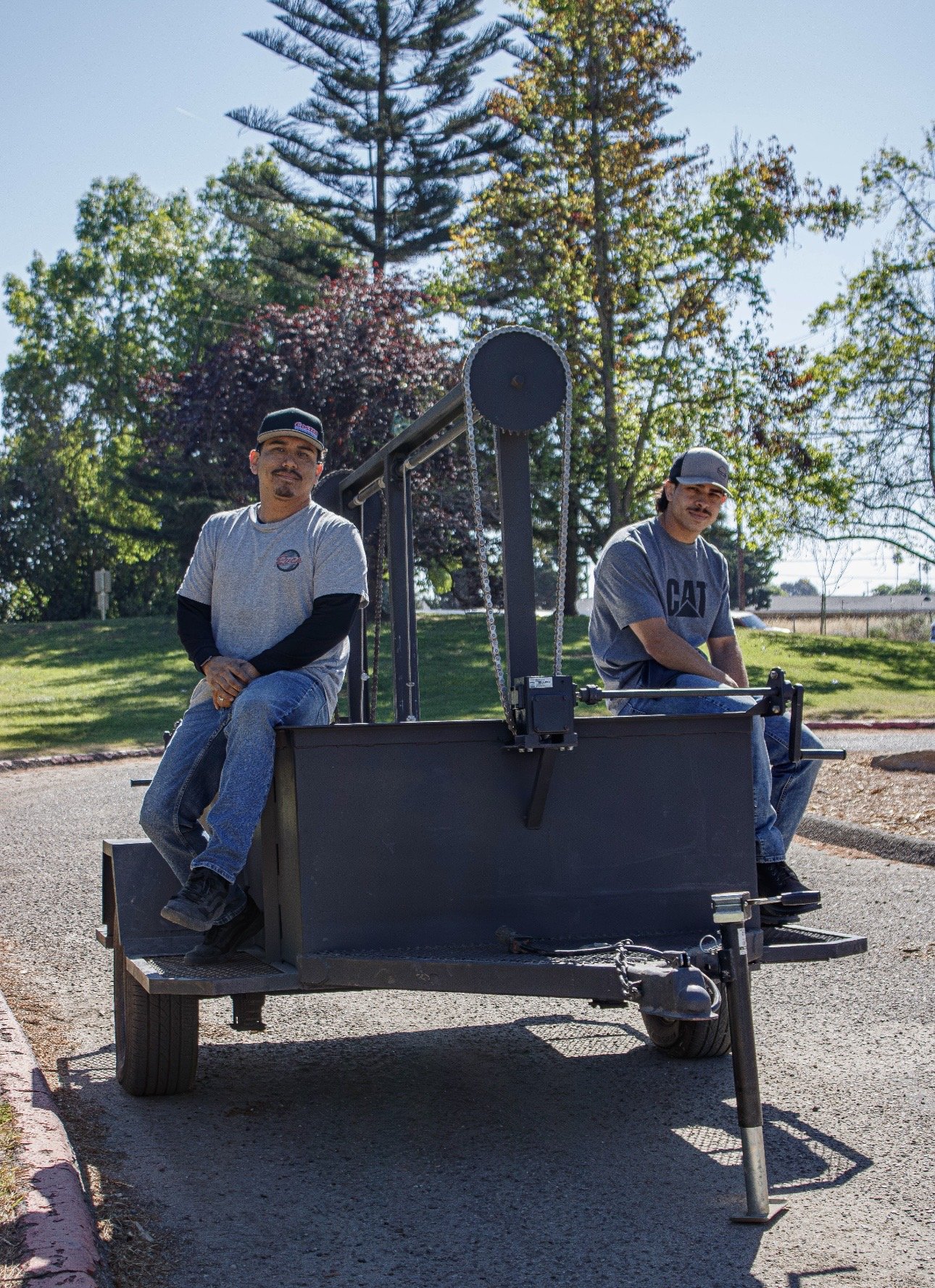Two men sitting on a black cart with a large circular saw blade attached, outdoors on a paved surface with trees and grass in the background.