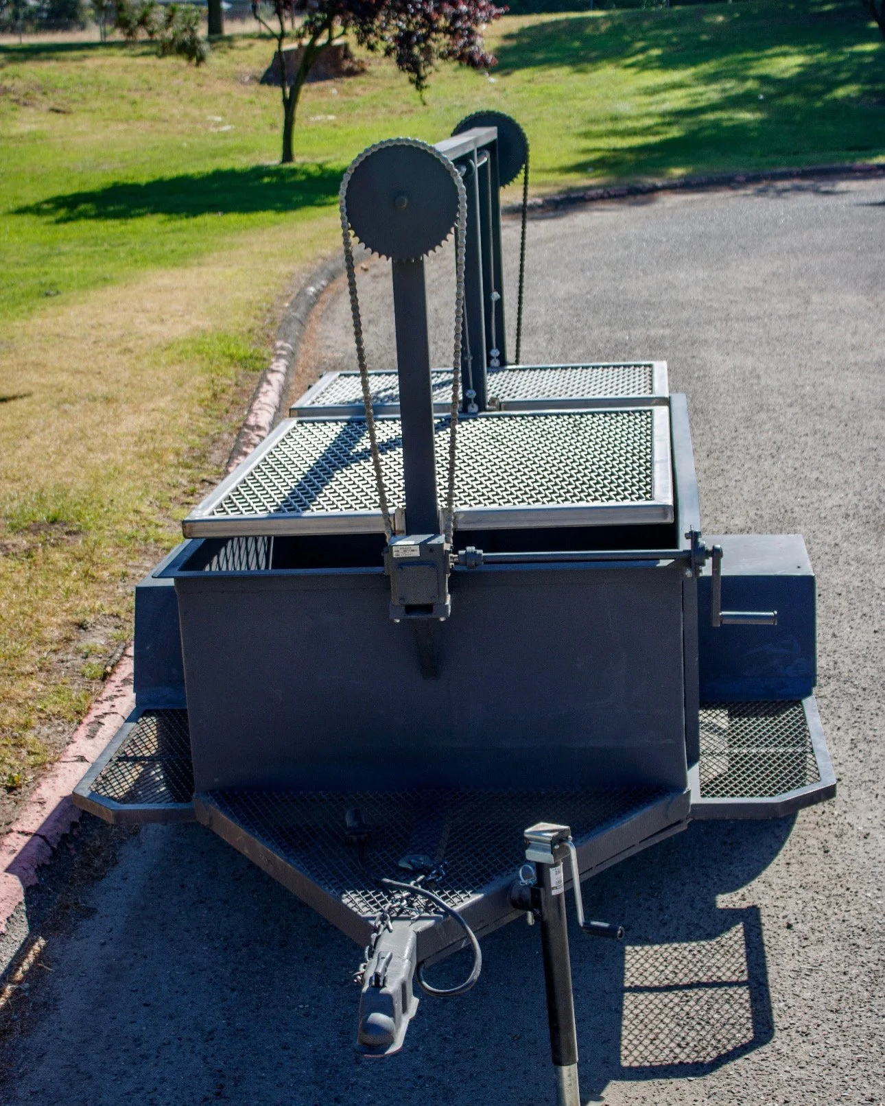 A mobile meat slicing machine with a large circular blade, chains, and conveyor surface situated outdoors on a parking lot with grass and trees in the background.