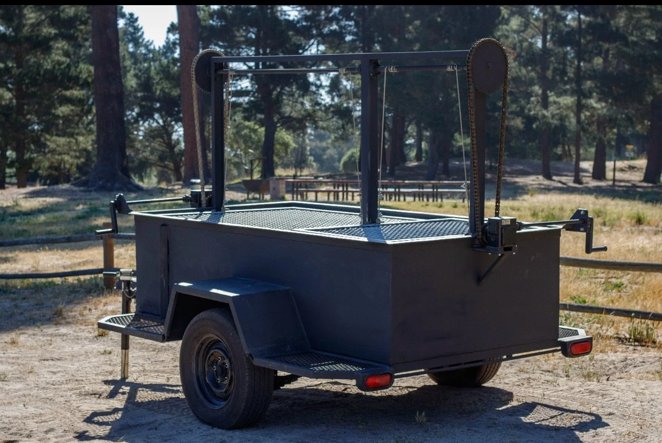 A black fishing or food truck trailer with a metal platform and support structures, parked on dirt in a wooded outdoor area.