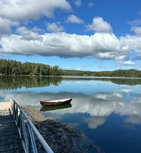 A calm lake with a small boat floating near the shore, surrounded by trees, under a partly cloudy sky.