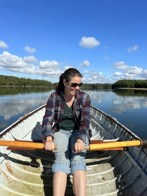 A woman in sunglasses and casual clothes sitting in a small boat on a calm river, surrounded by trees and blue sky with clouds.