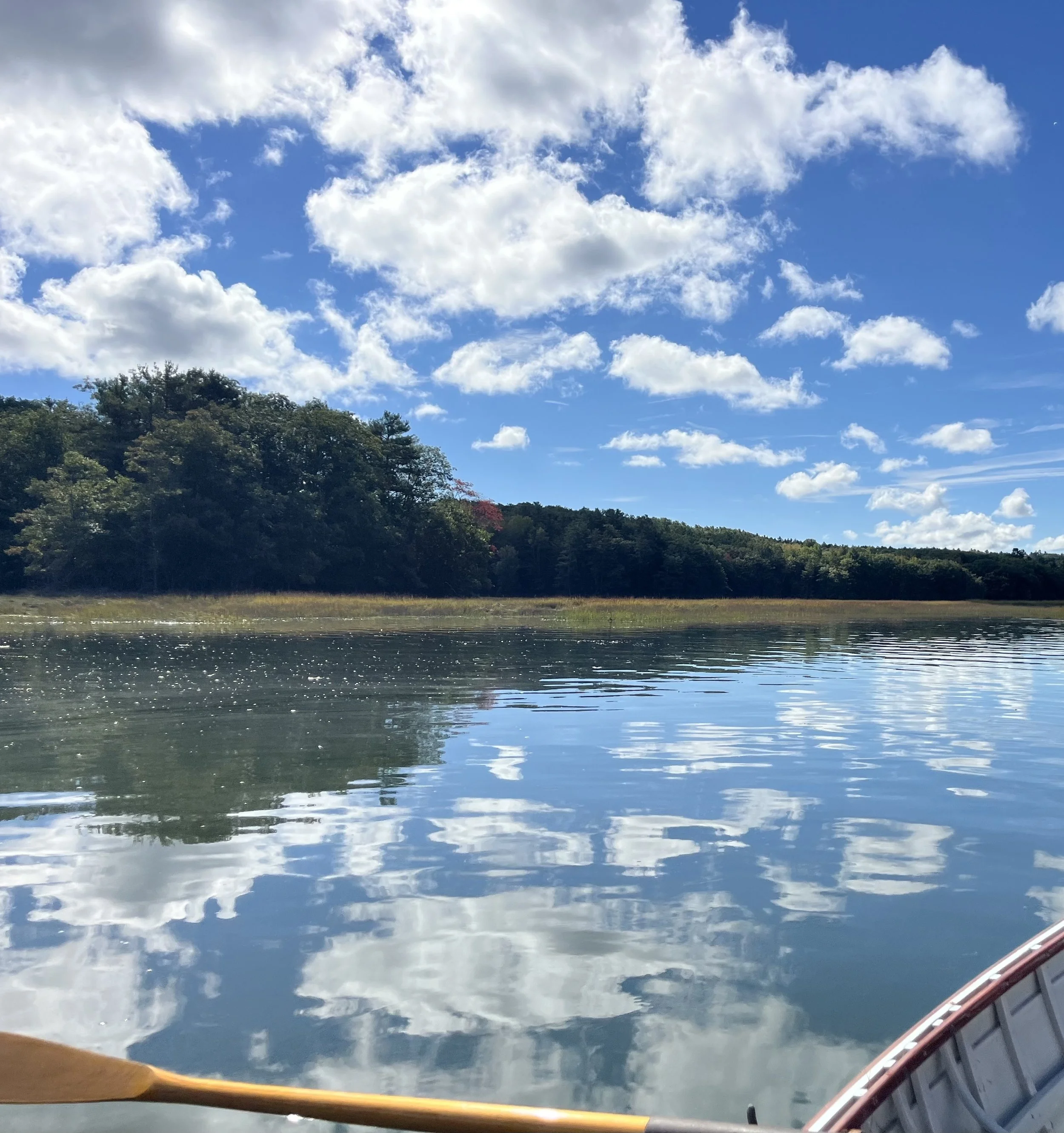 View of a bright blue sky with scattered white clouds, reflected on a calm body of water below. The edge of a wooden paddle and part of a boat's interior are visible at the bottom of the image.