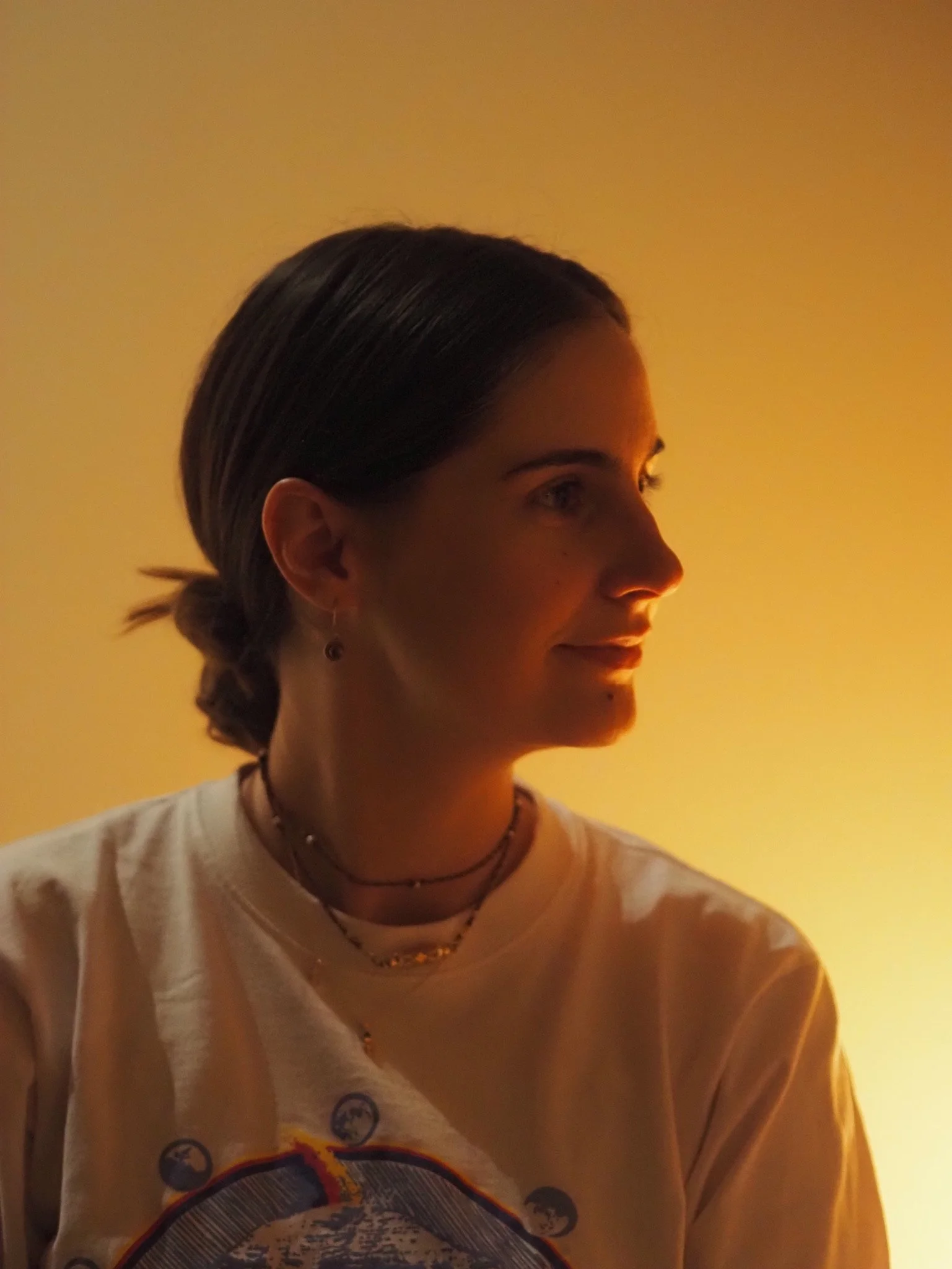 Side profile of a woman with dark hair tied back, wearing layered necklaces and a cream-colored T-shirt with a graphic design, against a warm yellow background.