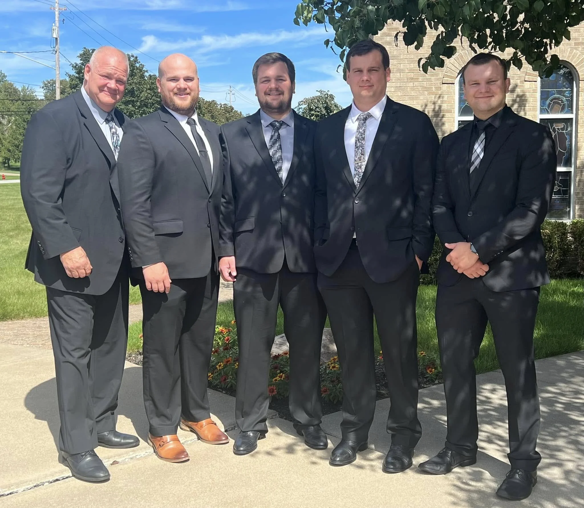 Six men in suits standing outdoors on a sidewalk, posing for a photo in front of a brick building and garden.