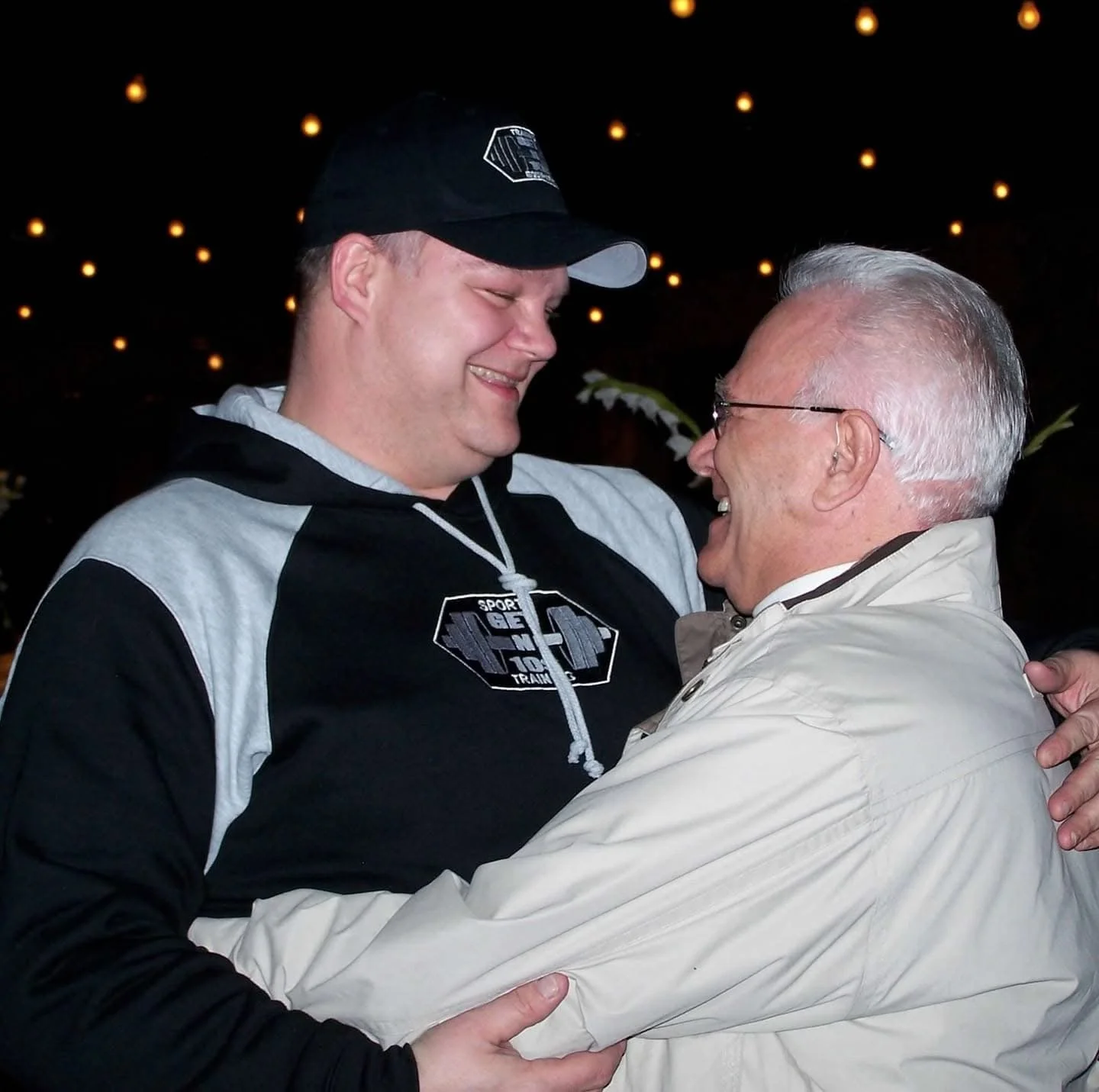 Two men smiling and embracing each other in a joyful hug, one young and the other older, in a dimly lit setting with warm lights in the background.