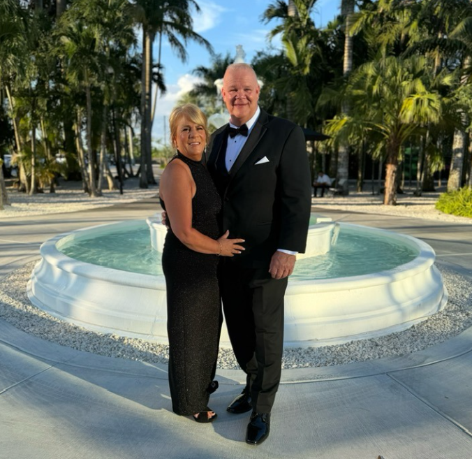 A smiling elderly couple in formal attire standing in front of a fountain surrounded by palm trees at a tropical outdoor venue.