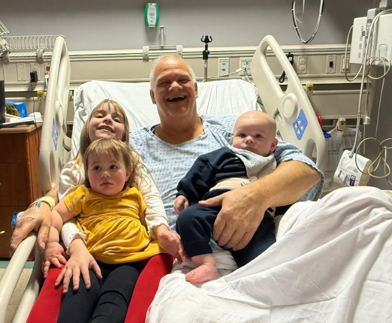A man in a hospital bed smiling with three children, two young girls and a baby, in a hospital room with medical equipment.