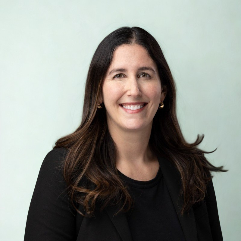Headshot of a woman with dark brown hair, wearing a black blazer and a black top, smiling against a plain light background.