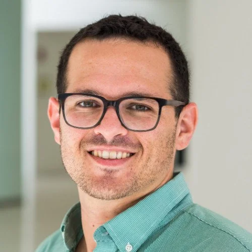 Close-up of a man wearing glasses and a green collared shirt, smiling in an indoor setting with blurred background