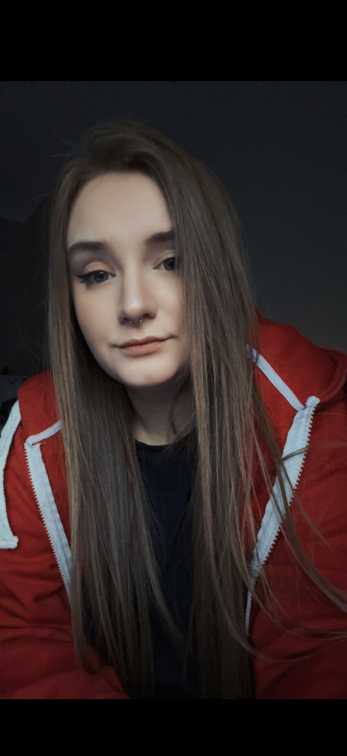 A young woman with long brown hair, wearing a red jacket with a white zipper and a black top, looking at the camera in a dimly lit room.