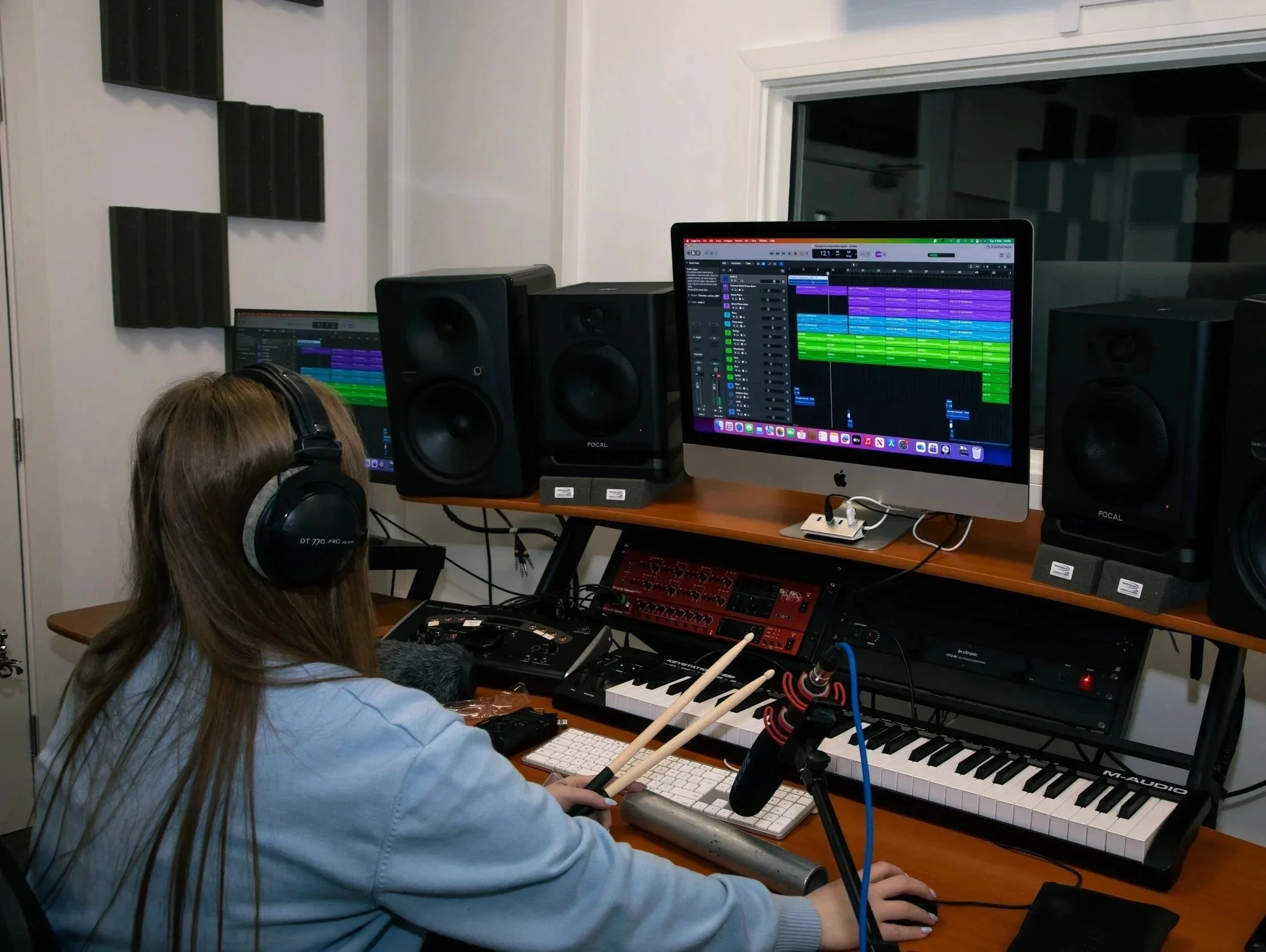 A person with long hair wearing headphones working on music production at a computer with multiple speakers and electronic music equipment in a recording studio.