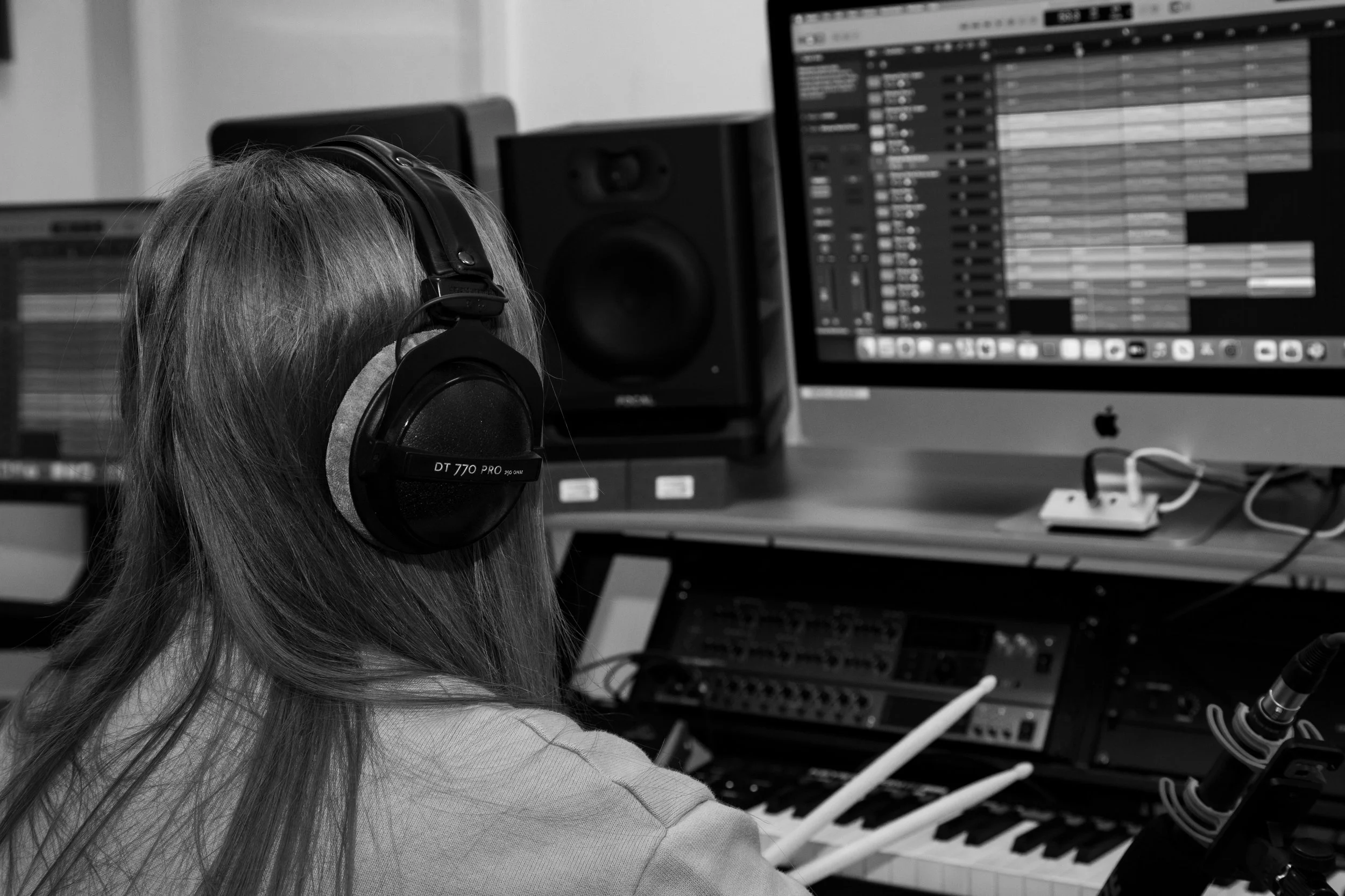 A person with long hair is wearing over-ear headphones and working on a digital audio workstation on a computer monitor, surrounded by audio equipment and musical instruments.
