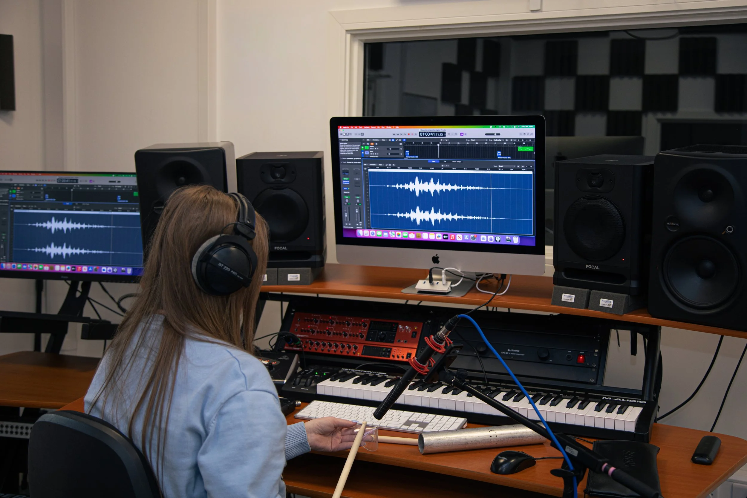 A person with long hair and headphones working on music production at a computer with audio editing software, surrounded by speakers, a keyboard, and recording equipment in a studio.