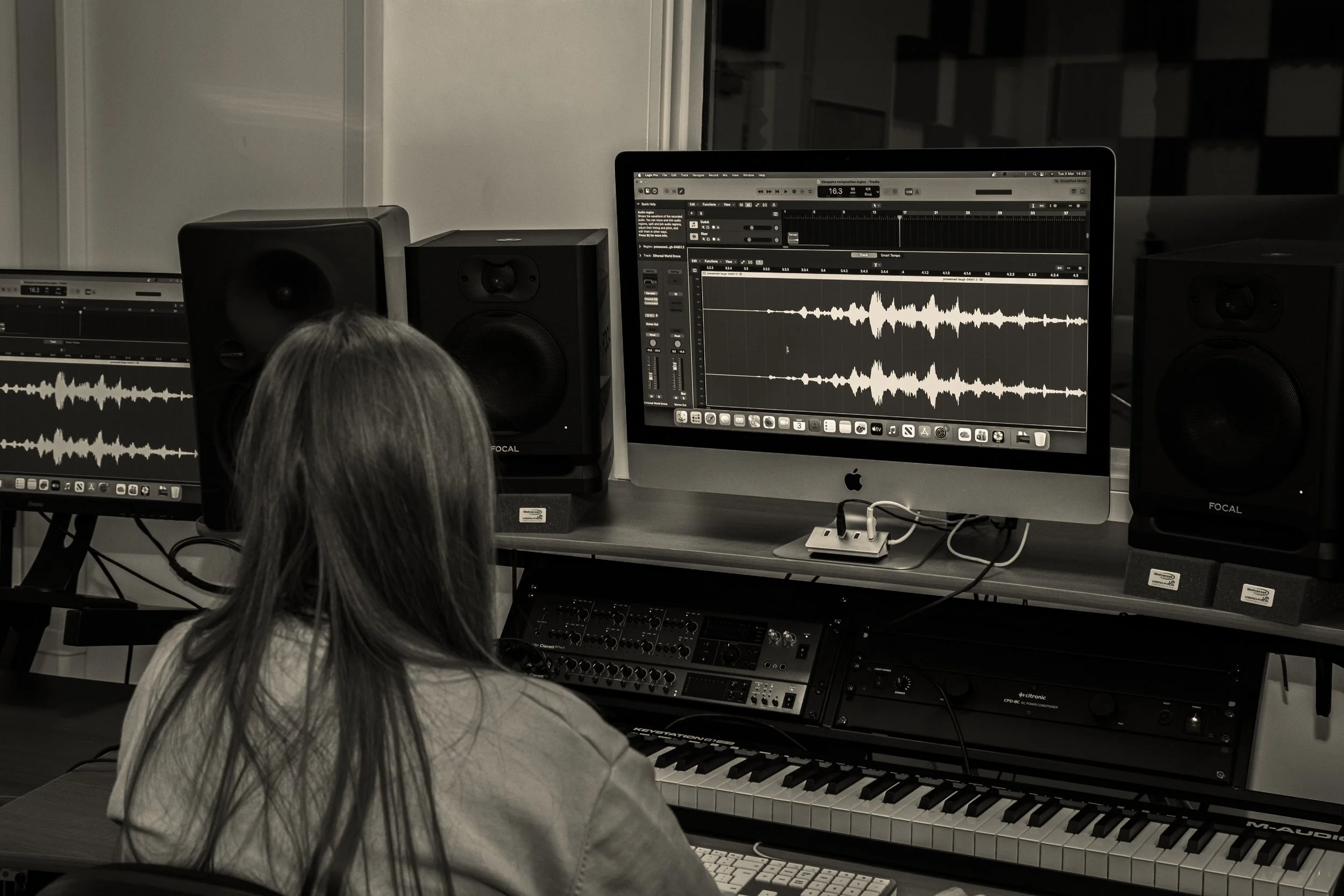 A person working on music production at a computer with audio editing software, surrounded by speakers and musical equipment.