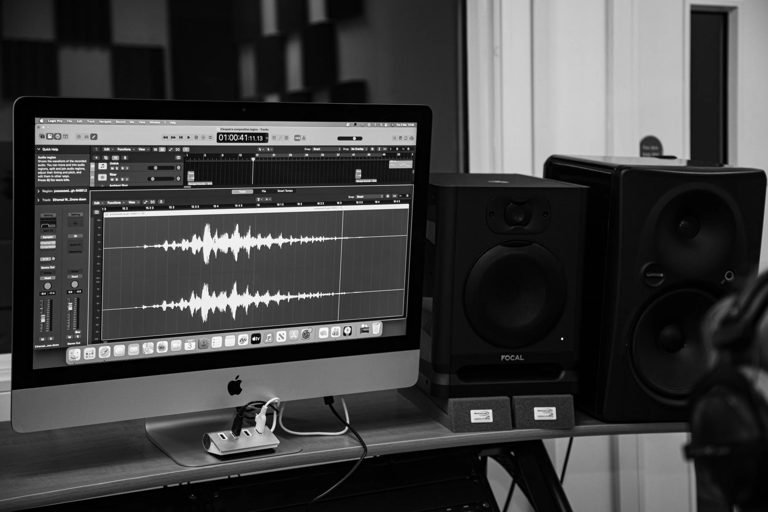 A computer monitor displaying audio editing software with a waveform, studio speakers, and a USB hub on a wooden desk.