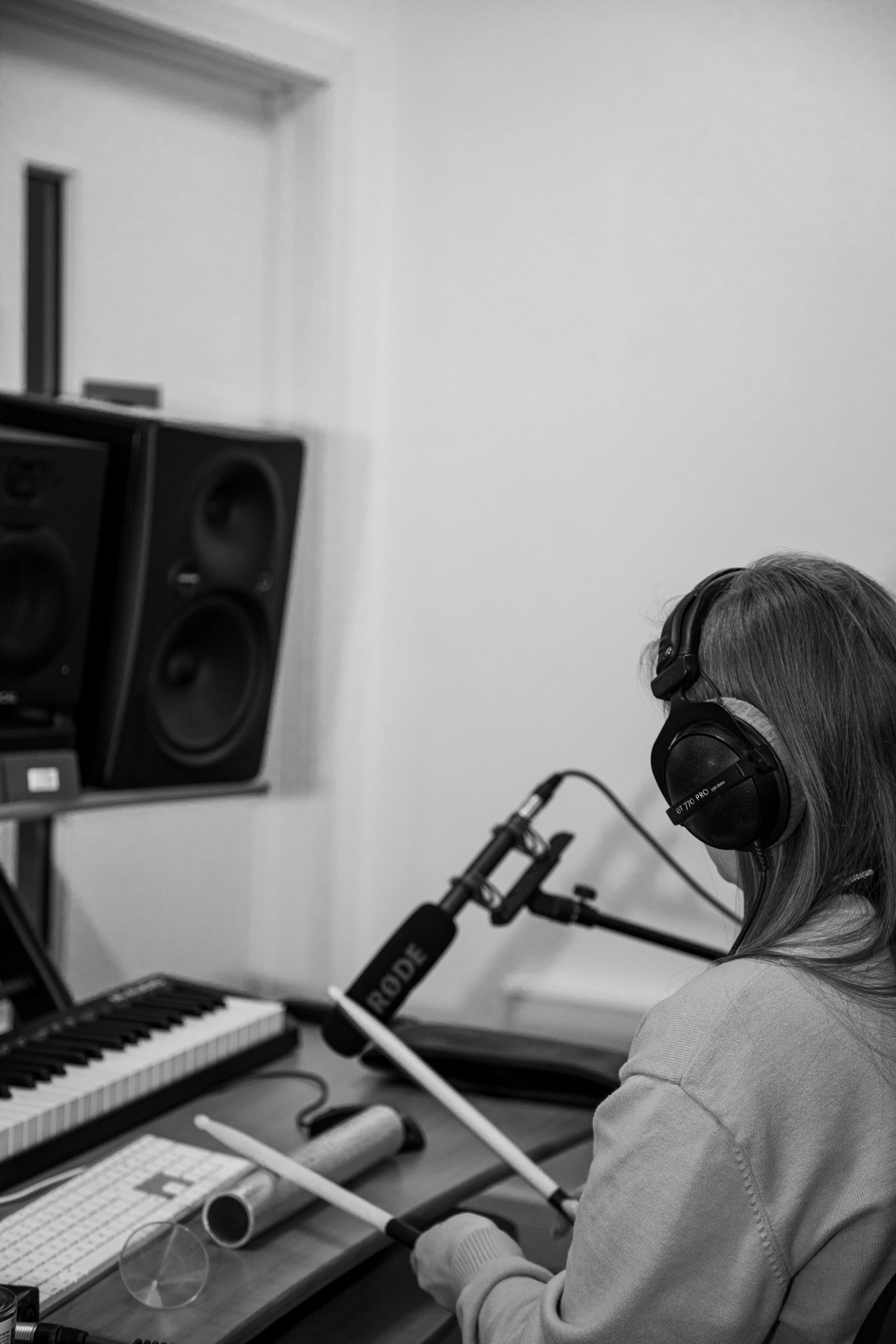 A woman with gray hair, wearing headphones, is seated at a desk recording into a microphone with a pop filter. In front of her is a keyboard, a computer keyboard, and various music production equipment, in a studio or recording space.
