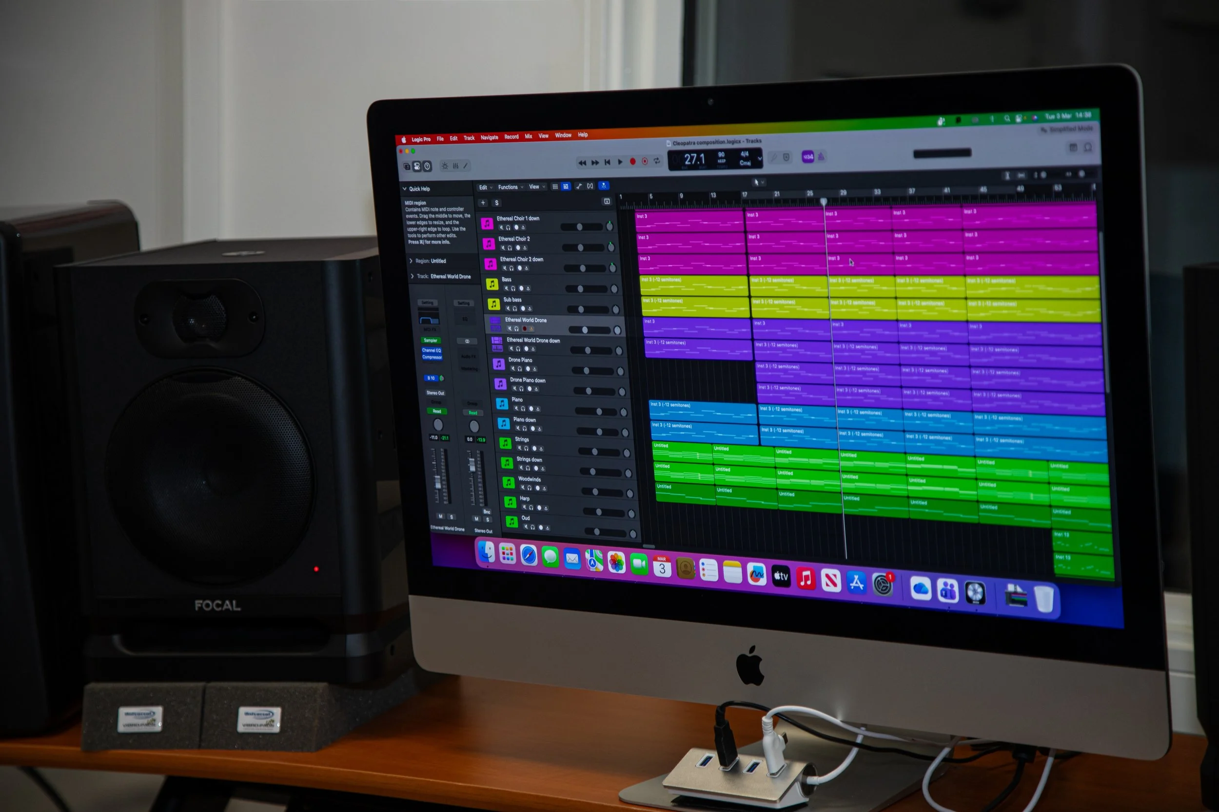 A computer monitor displaying digital music production software with colorful tracks, a speaker to the left, and power strips with connected cables on a wooden desk.