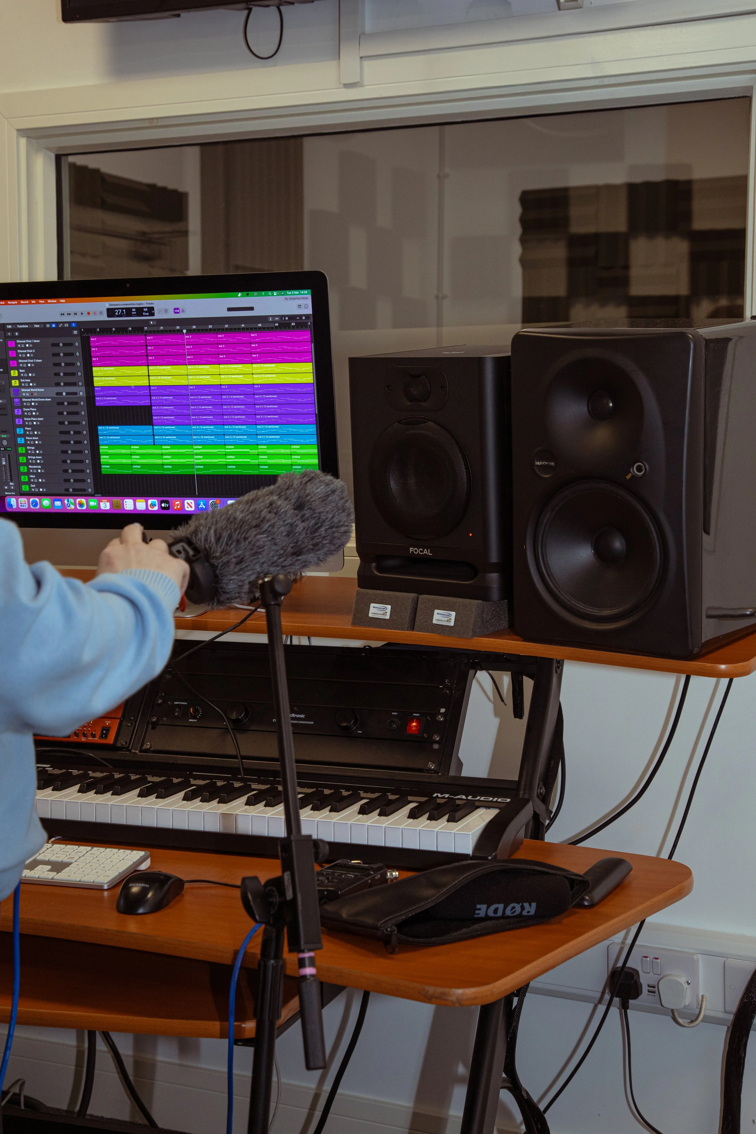 Music production setup with a keyboard, computer monitor displaying a colorful digital audio workstation, studio speakers, and audio equipment on a wooden desk in a recording studio.