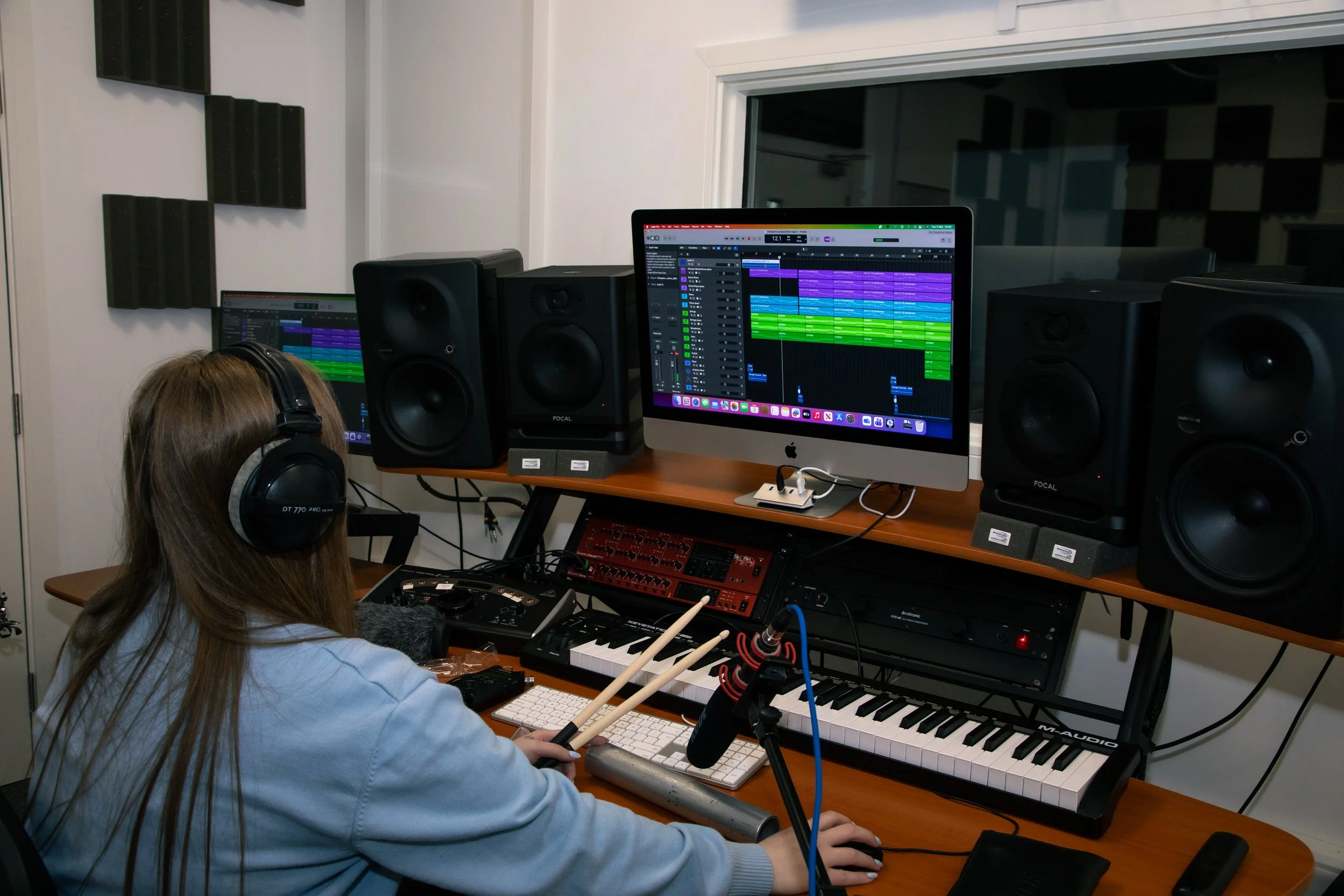 A person in a recording studio wearing headphones, playing digital drums, and viewing music production software on a computer screen. The room has soundproofing foam on the walls.