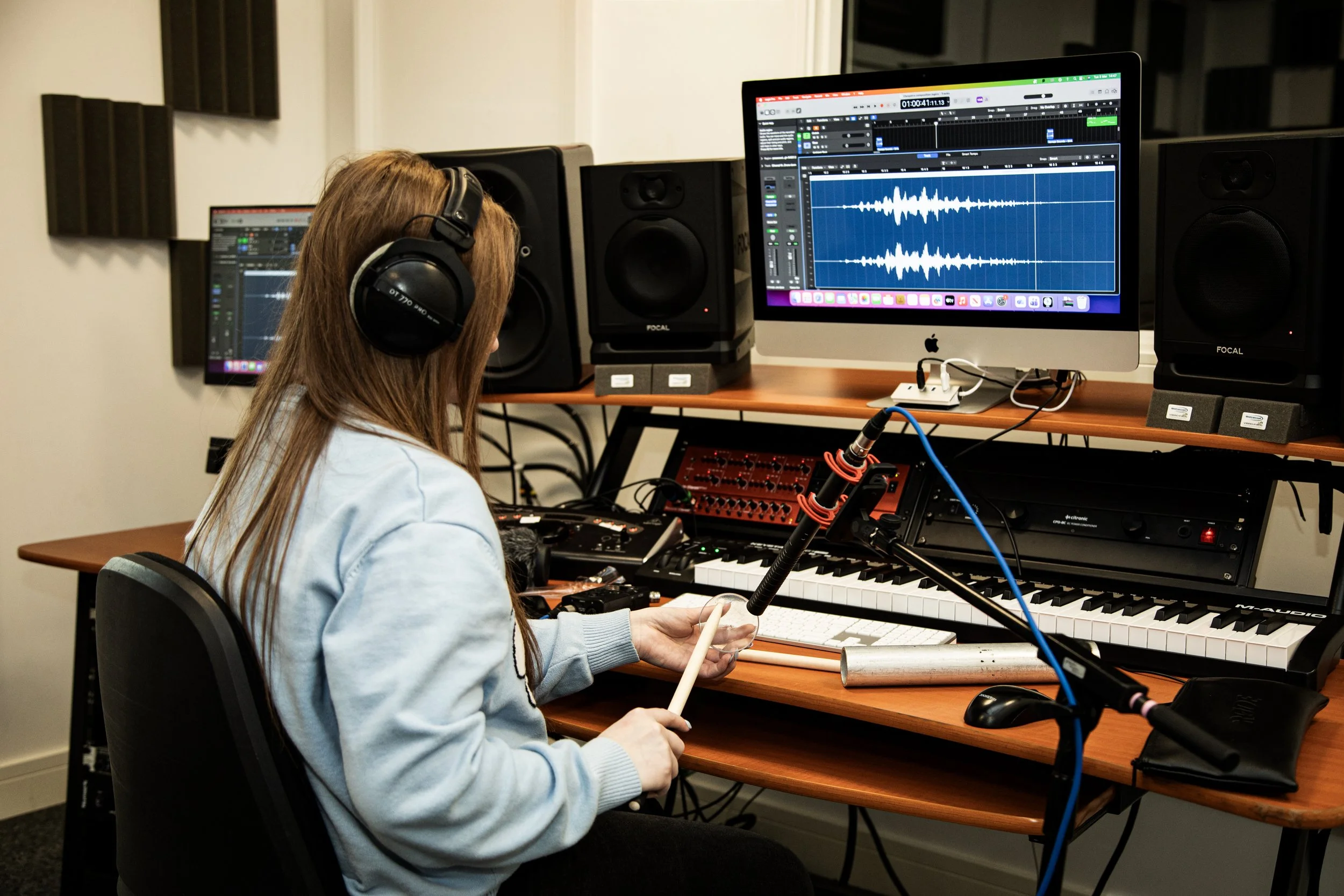 A woman with long hair wearing headphones and a light-colored sweater sitting at a desk in a recording studio with audio equipment, a computer displaying audio editing software, and speakers.