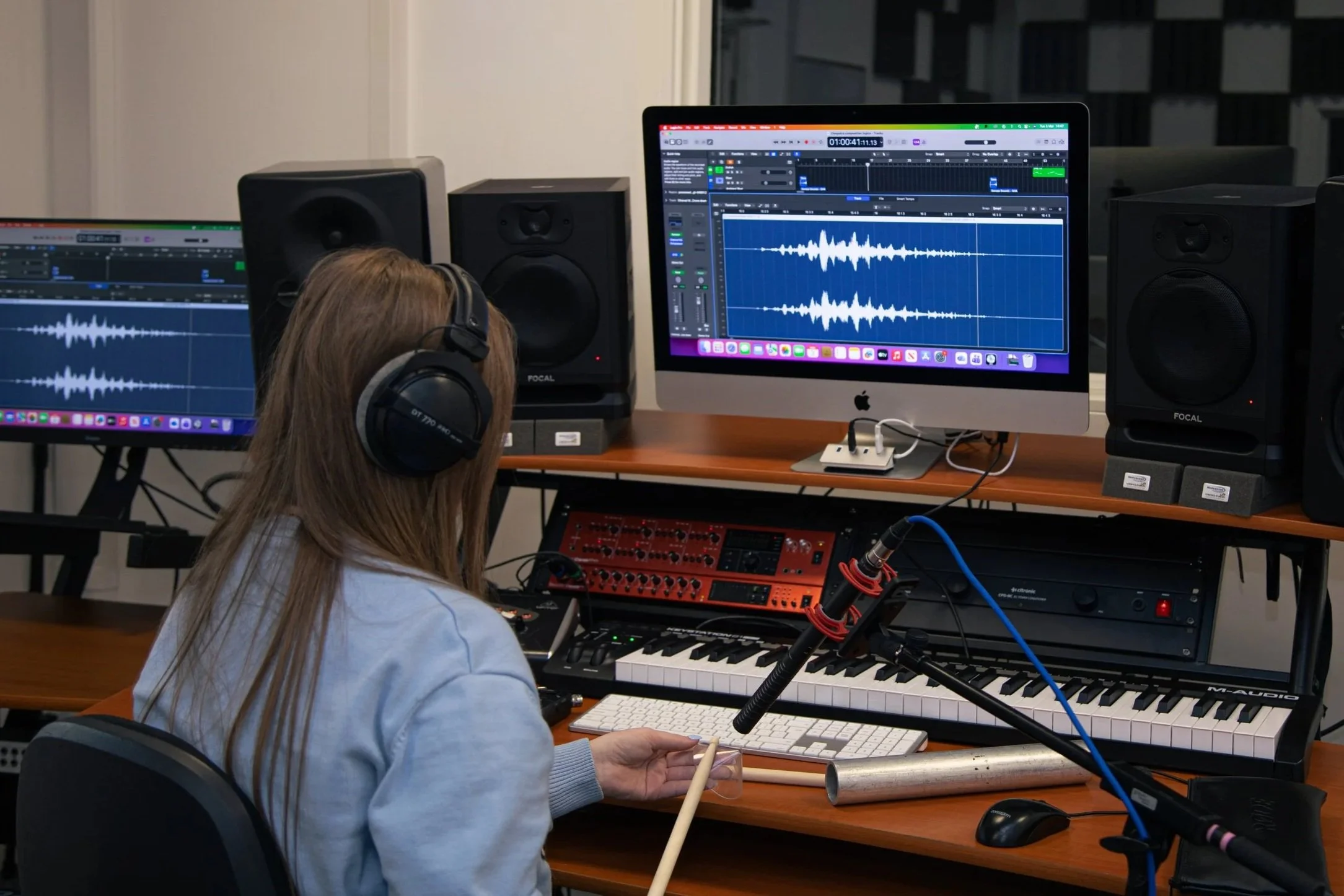 A woman wearing headphones working on music production at a computer with audio editing software displayed on the screen, surrounded by studio monitors, MIDI keyboard, and audio equipment.