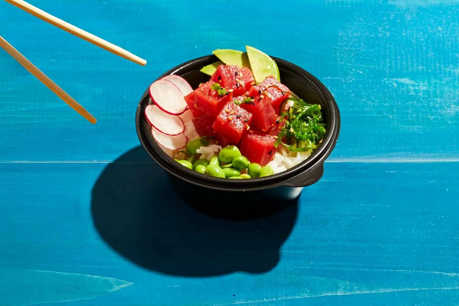 Bowl of poke with cubes of raw tuna, sliced radishes, avocado, seaweed salad, edamame, and rice on a blue wooden table