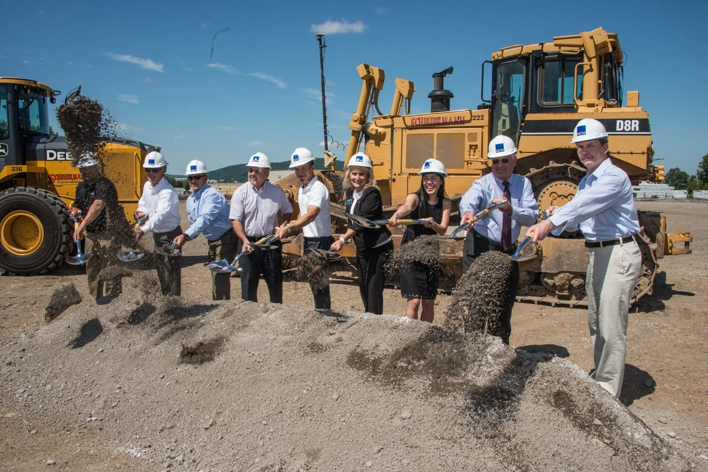 Group of people wearing construction helmets and formal attire doing a groundbreaking ceremony with shovels in front of construction machinery on a clear day.