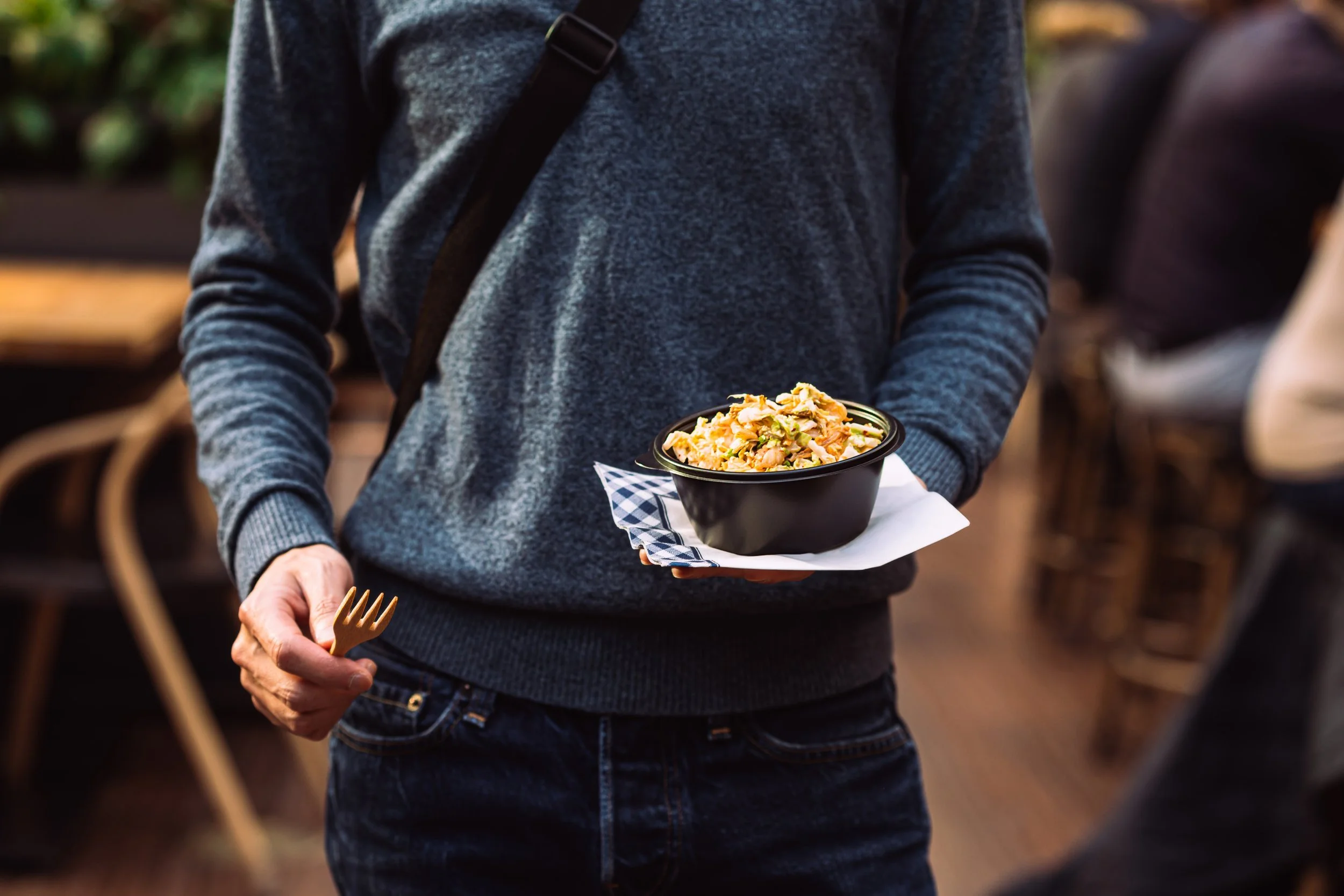 Person in a gray sweater holding a bowl of food with a paper napkin under it, standing in a restaurant or café.