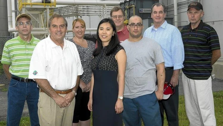 Group of eight people standing outdoors in front of industrial equipment, smiling at the camera.