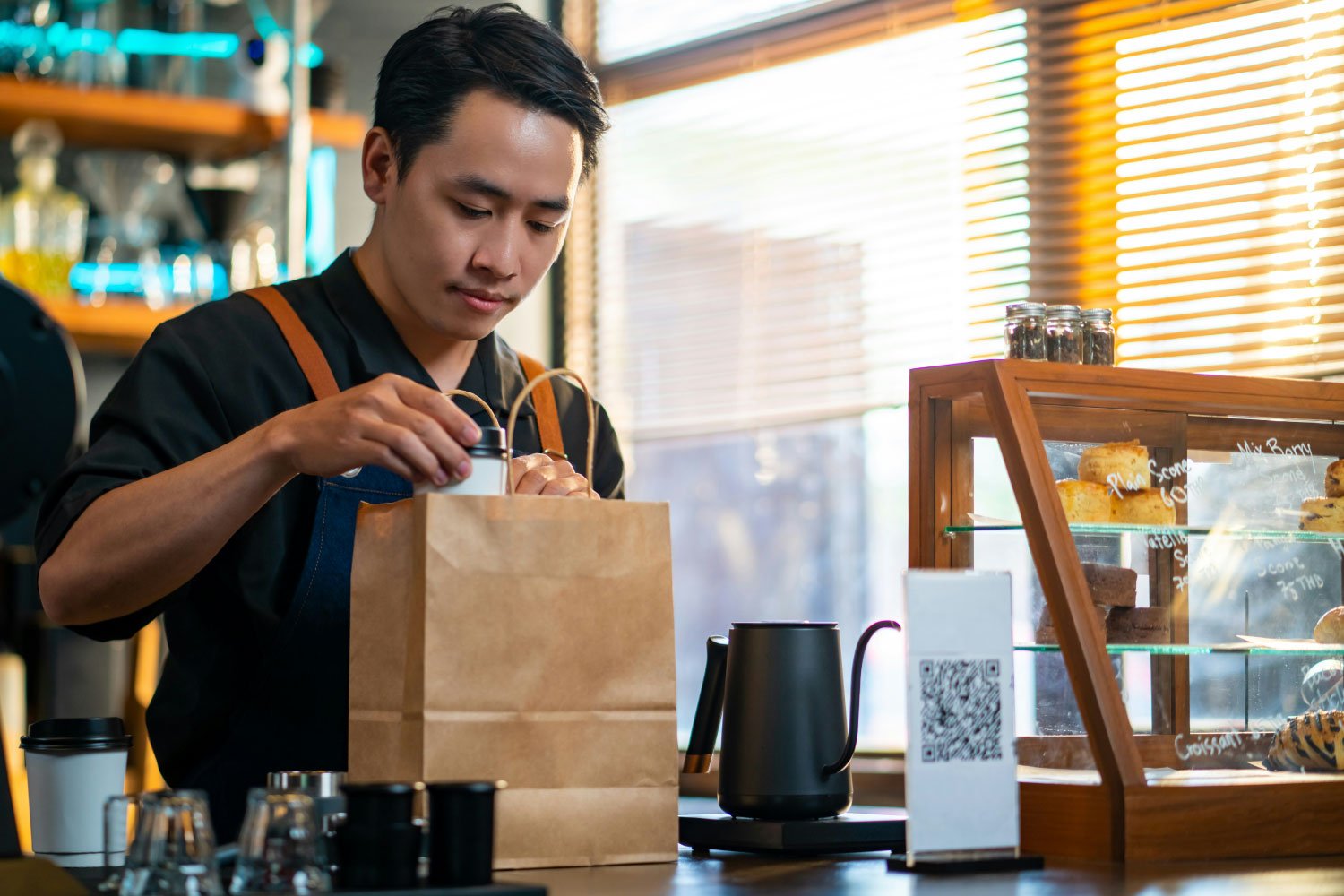 A young man packing a brown paper bag at a cafe counter, with pastries displayed in a glass case to his right and coffee cups in front of him.